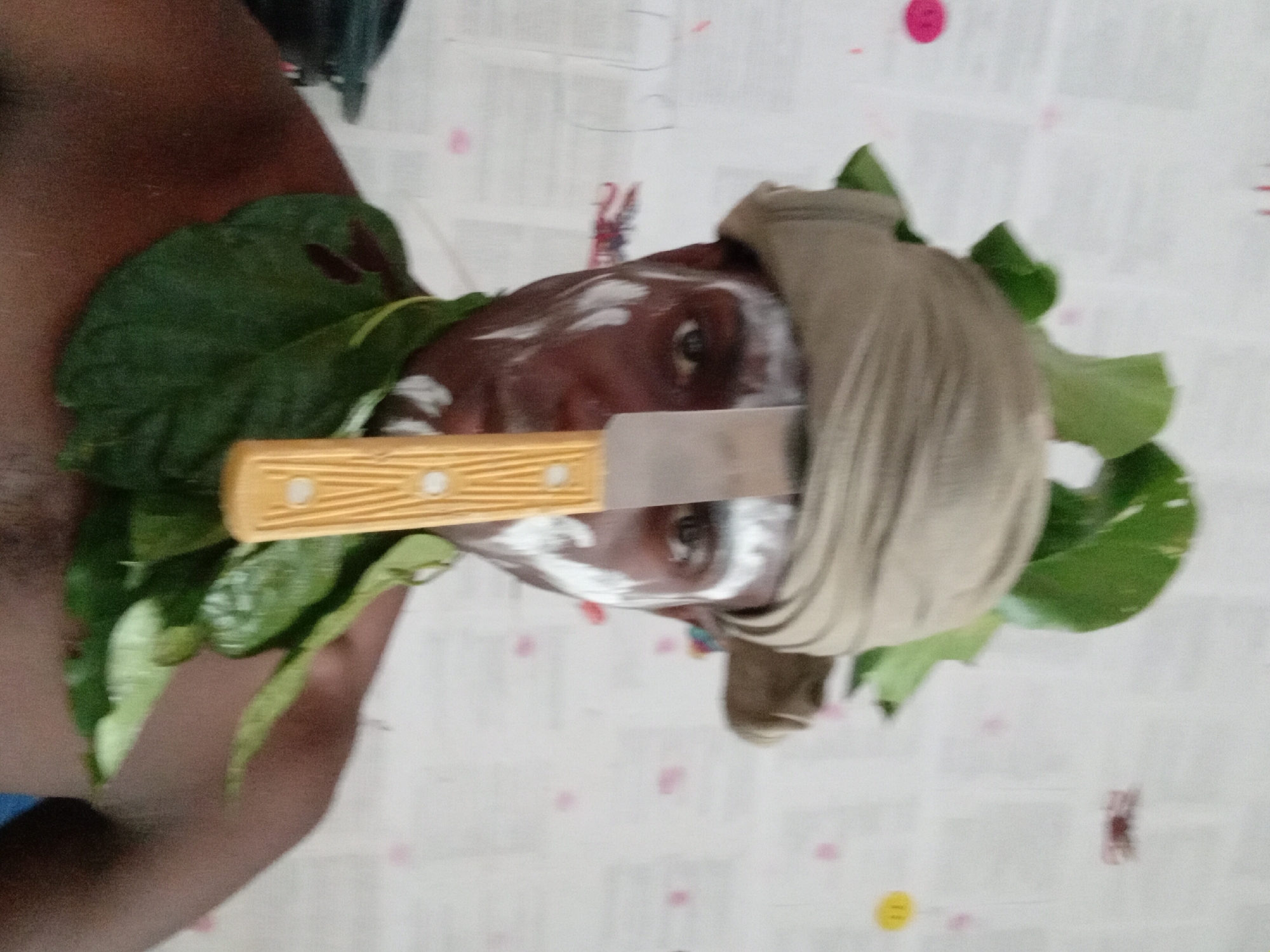 A man with a dark complexion, partially covered in white face paint, is the central subject. He is adorned with large green leaves around his head and neck. A beige-colored cloth is wrapped around his head, partially obscuring his forehead. A knife with a yellow handle is positioned horizontally across his face, held between his eyes. The background consists of a light-colored patterned surface. The scene appears to be set indoors. The location is Akwete, Nigeria.