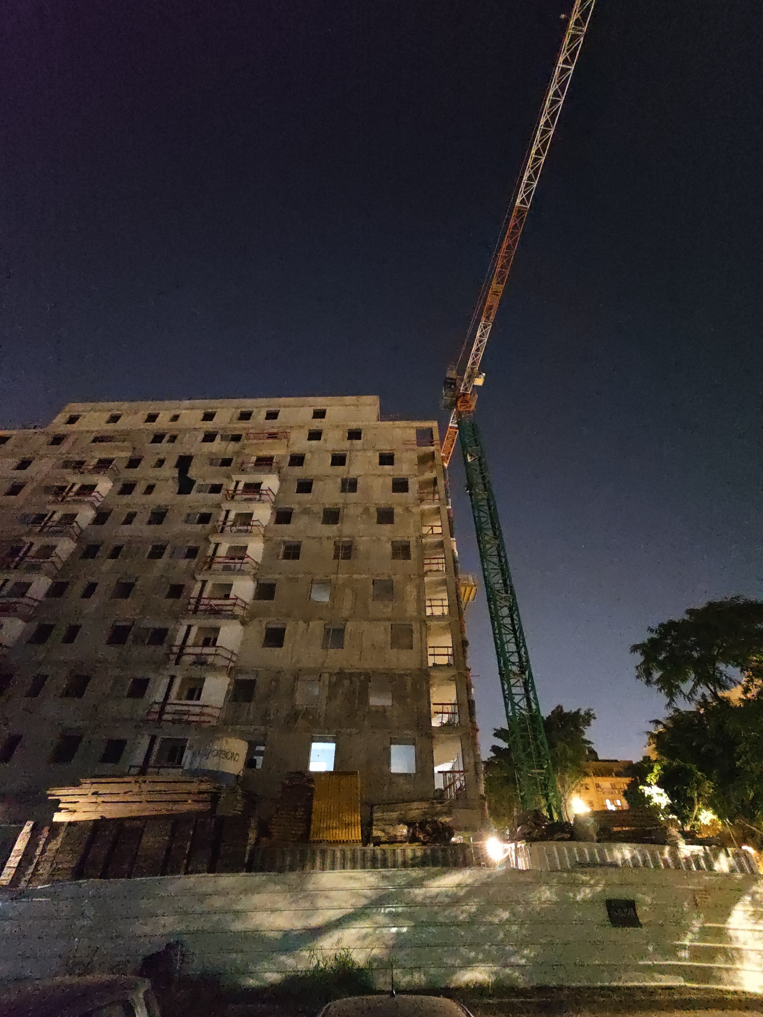 An outdoor, low-angle, night-time shot captures a tall construction crane reaching diagonally upwards against a dark, starry sky, with its boom section displaying alternating orange and white segments. The crane is positioned next to a large, unfinished concrete building that appears to be under construction. The building has multiple floors with numerous rectangular windows, many of which are dark, but some on lower floors are illuminated. Several balconies on the building are incomplete, featuring exposed concrete and red railings. At the base of the building, stacks of wooden planks and other construction materials are visible. In the foreground, a textured concrete barrier or wall is present, with scattered patches of grass and shadows cast from the building and crane. To the right of the building, a cluster of trees and some illuminated buildings are visible in the background, suggesting an urban environment. The image appears to be taken in Petah Tikva, Israel, a city known for its ongoing urban development and construction projects.