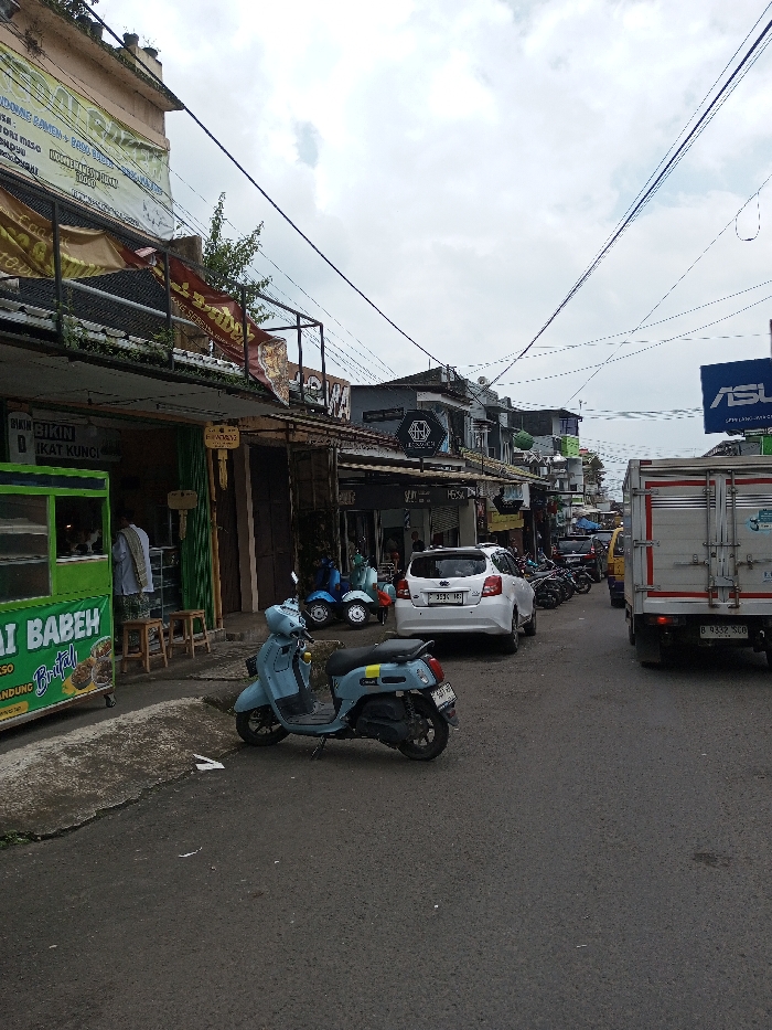The image displays an urban street scene under an overcast sky. The street is asphalt, flanked by multi-story commercial buildings on both sides.
On the left side, in the foreground, a light-green food cart is visible, bearing text such as "MIE AYAM BABEH" and "BANDUNG BAKSO." A person in a light-colored garment stands behind the cart, facing away, while another individual is partially discernible inside the cart. Two wooden stools are positioned near a shop entrance to the right of the food cart. Above these, building facades display various signs and banners, including one stating "BIKIN KUNCI" and another featuring "MIE AYAM" and "MISO" text. A light blue scooter with yellow accents and an Indonesian license plate is parked on the street in the immediate foreground. Further down the left side, a white hatchback car with an Indonesian license plate "F 3536 HS" and several other motorcycles, including two light blue Vespa-style scooters, are parked along the curb.
On the right side of the street, the rear of a white box truck with red striping and an Indonesian license plate "B 9332 SLO" is prominent in the foreground. Above the truck, a blue sign featuring the "ASUS" logo is mounted on a building. Additional commercial buildings extend into the background, one displaying a black octagonal logo sign. Numerous overhead power lines and communication cables are visible against the cloudy sky, crisscrossing above the street. The street continues into the distance, populated with more parked vehicles and buildings.