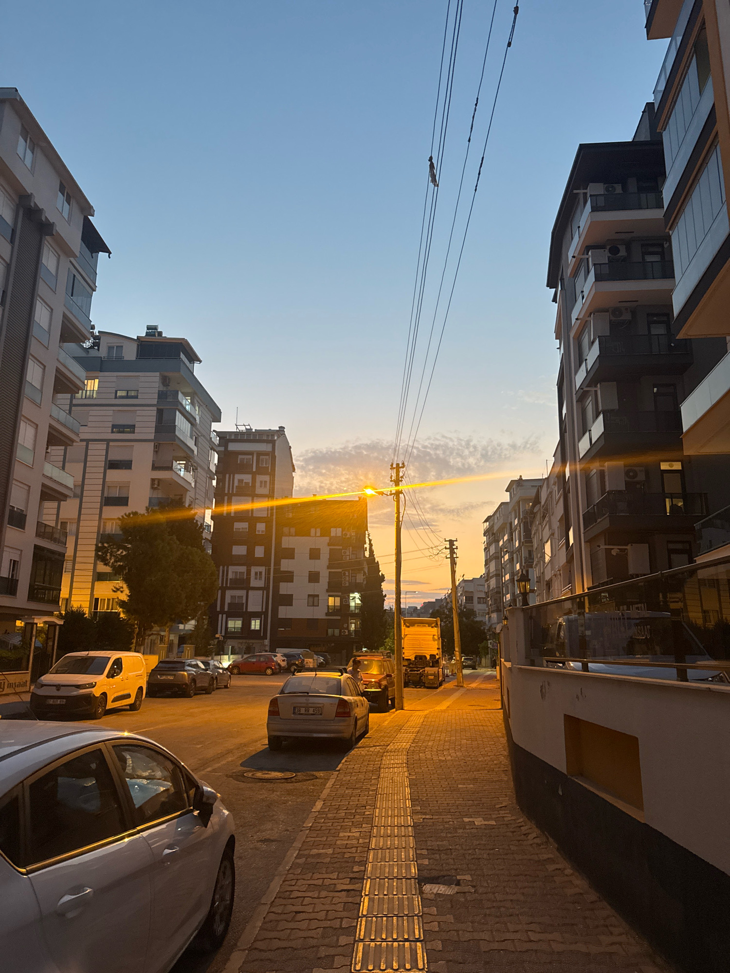 The image depicts a street scene in Antalya, Turkey, during sunset. The sky is a gradient of blue and orange, with scattered clouds. Tall, modern apartment buildings line both sides of the street, featuring balconies, air conditioning units, and numerous windows. Power lines are visible overhead, crisscrossing the sky.
Several cars are parked along the street, including a white van, a silver sedan, and a darker colored car. A yellow truck is also present further down the road. The street is paved with interlocking bricks, and a textured walkway, likely for the visually impaired, runs alongside it. Streetlights cast a warm, orange glow on the pavement and buildings, enhancing the twilight atmosphere. The overall impression is of an urban residential area at dusk.