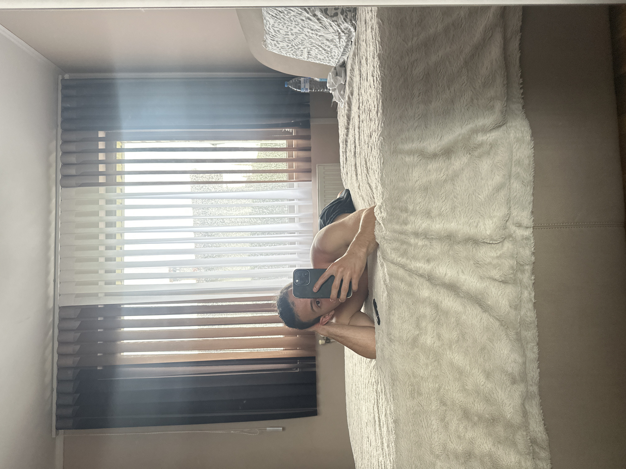 The image depicts an indoor scene in a bedroom located in the city of Saraia, Bulgaria. The photograph is a selfie taken in a mirror by a person, appearing male, lying prone on a large bed covered with a light-colored, textured faux fur or fleece blanket. The individual's torso is bare, and dark shorts are visible. Their left arm supports their head, while their right hand holds a dark-colored smartphone, obscuring most of their face except for their left eye, nose, and forehead. A black pen-like object lies on the bed in front of them.
Behind the bed, a large window spans the wall, fitted with vertical blinds or curtains. These coverings transition in color from dark brown/black on the left, to medium brown/tan in the center, and white/translucent on the right. Bright sunlight streams through the partially open blinds, creating strong highlights and a subtle lens flare in the upper right quadrant of the image.
To the right of the person, a light beige headboard is visible, against which a pillow with a grey and white patterned case rests. A clear plastic water bottle with a blue cap and a crumpled pile of white fabric are positioned on the bed near the headboard. A white radiator is visible below the window on the far right. The walls are a light, possibly beige or pinkish-beige, and the ceiling is white with molding.
