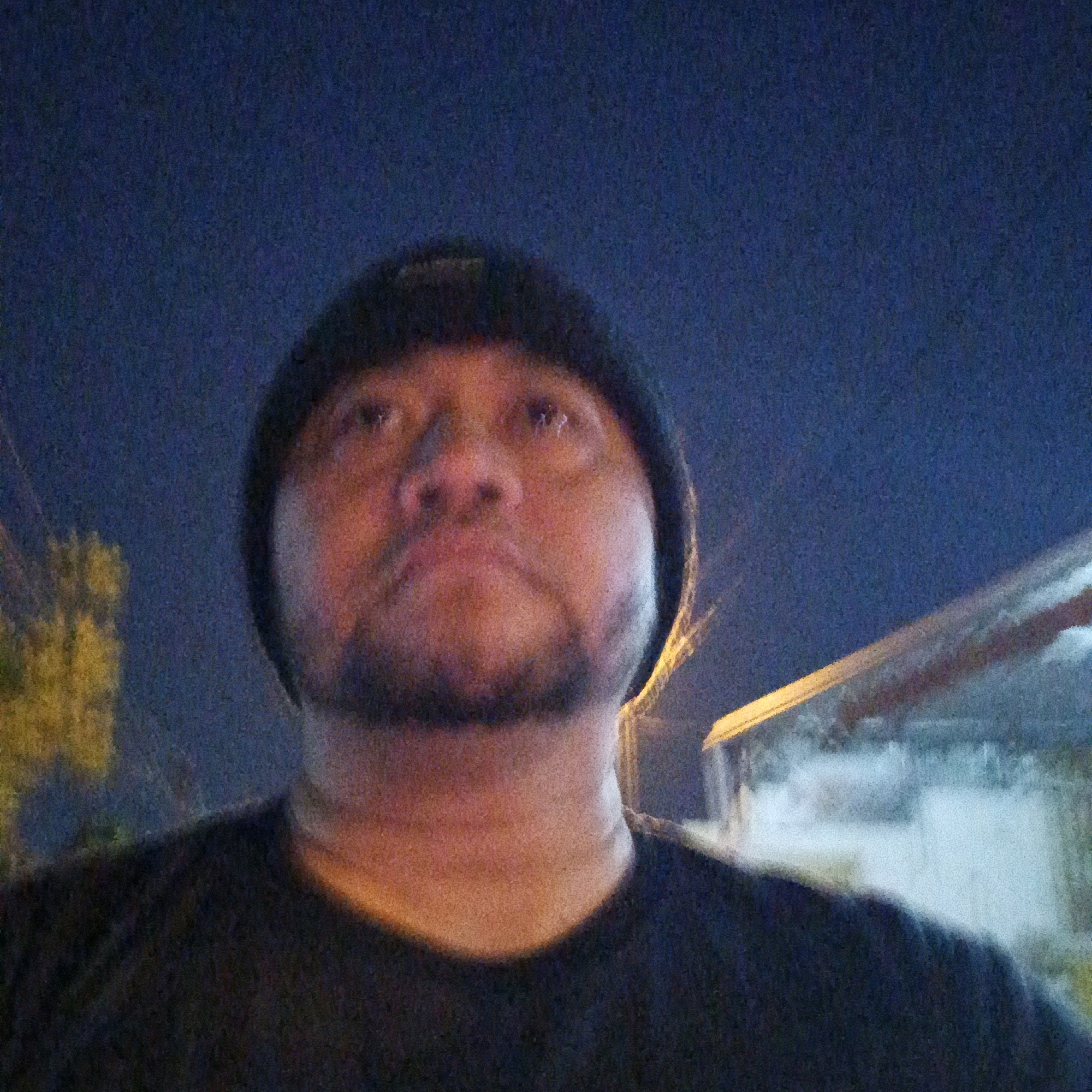 The image is a low-light, selfie-style photograph taken outdoors at night in Salatiga, Indonesia. The primary subject is a person wearing a dark beanie and a dark t-shirt. The person's face is turned upwards and slightly to the left, with their gaze directed upwards. They have a short beard and mustache.
In the background, the night sky is visible, appearing dark blue. To the left of the subject, there are blurred yellow-green shapes that suggest foliage or trees illuminated by artificial light. To the right and in the background, there is a building or structure with a sloping roofline that is illuminated by warm-toned lights. Some white smoke or mist is visible near this structure. The overall image has a grainy texture due to the low light conditions.