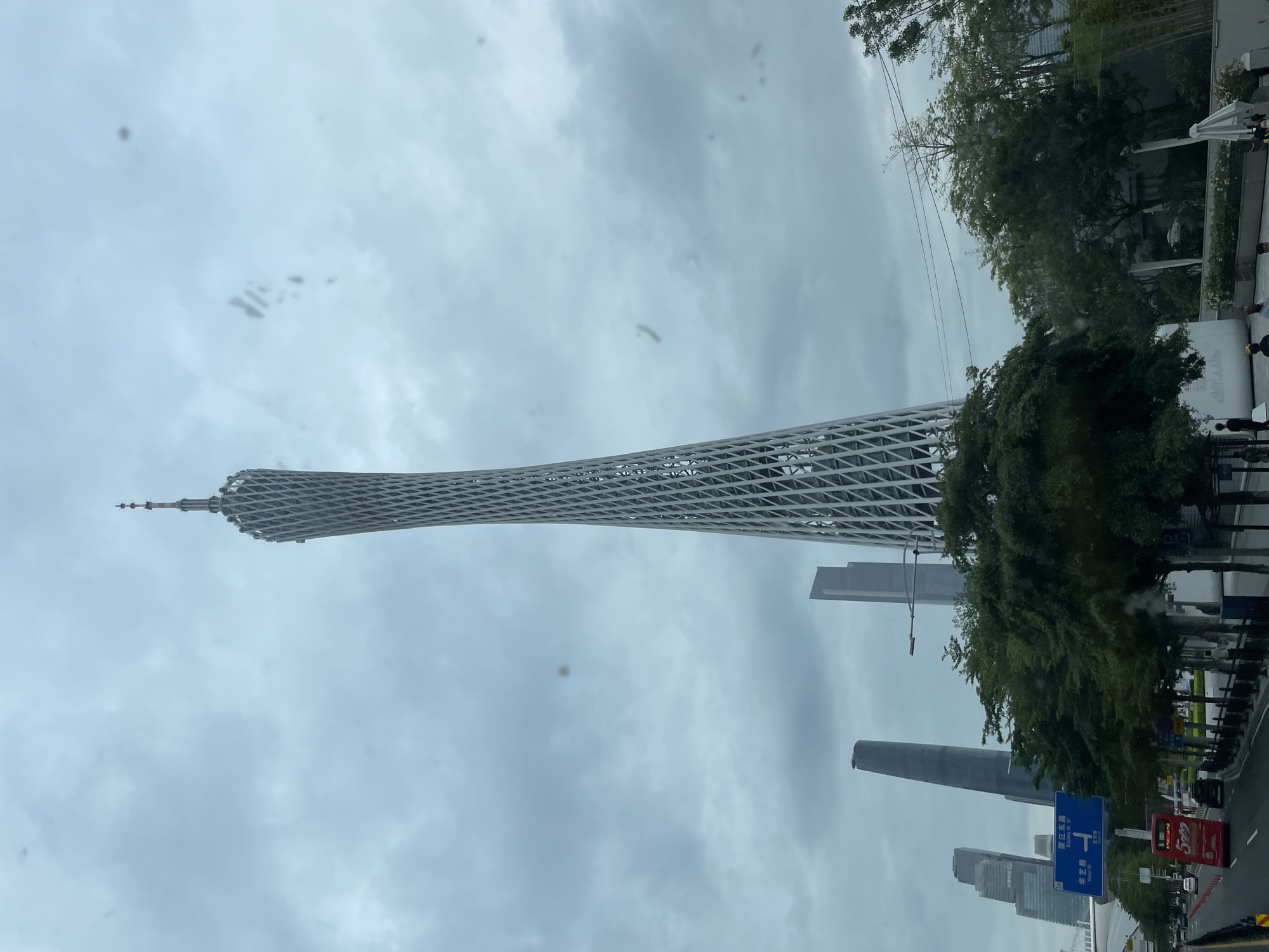 The image shows the Canton Tower in Haizhu District, Guangzhou, China. The tower is a prominent structure dominating the skyline under a cloudy sky. There are buildings next to the tower, and trees and foliage are seen along with the road traffic below.