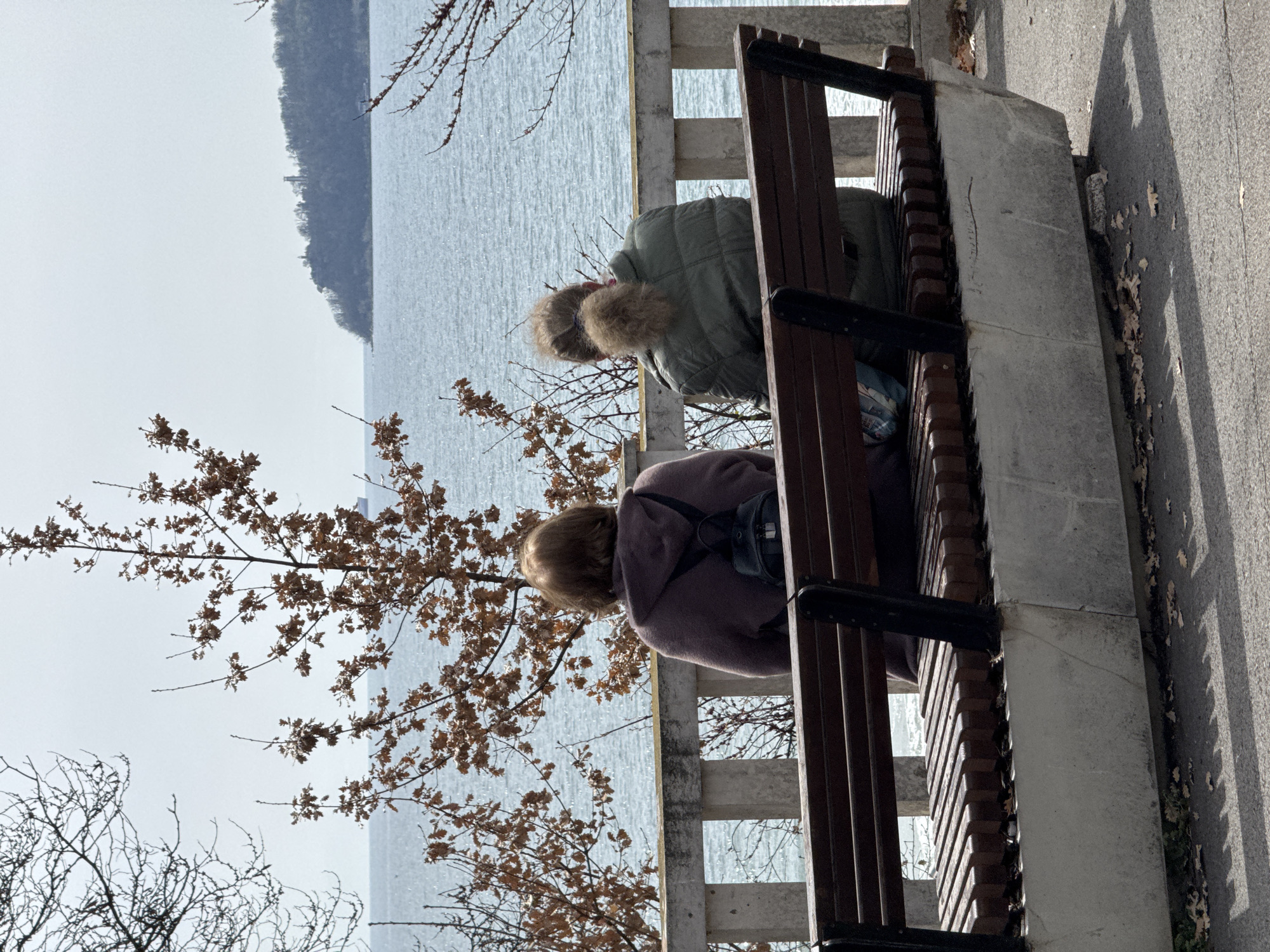 Two women are sitting on a park bench facing the sea, likely in Varna, Bulgaria. The bench has a dark wood and black metal frame with a concrete base. One woman is wearing a green jacket with a fur-trimmed hood, and the other is wearing a purple jacket with a black backpack. Bare tree branches frame the left side of the image. The sea appears calm and reflects the sky, with land visible in the distance. A paved walkway runs along the right side of the bench, strewn with fallen leaves. A concrete fence is behind the bench. The image is rotated clockwise, making the bench appear vertical.