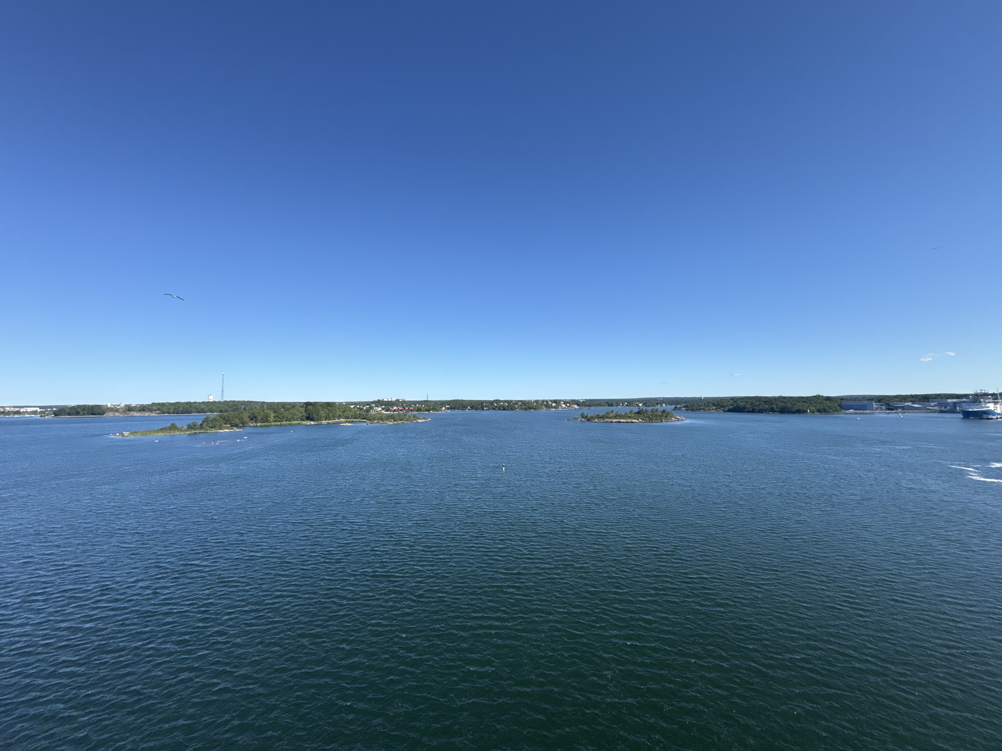 The image captures a wide view of a body of water with several islands dotting the horizon, set against a clear blue sky. The water is calm with gentle ripples, reflecting the bright sunlight. Lush green trees cover the islands, and some small structures, possibly houses or buildings, are visible on the mainland in the distance. A tall tower with an antenna stands prominently on one of the islands. In the far right of the image, a large blue and white ship is partially visible, moving away from the viewer. A single bird is seen flying in the upper left quadrant of the sky. The scene suggests a tranquil day in a coastal or archipelago setting. This view is consistent with the archipelago surrounding Karlskrona, Sweden, a city known for its naval history and its location within a scenic coastal landscape.