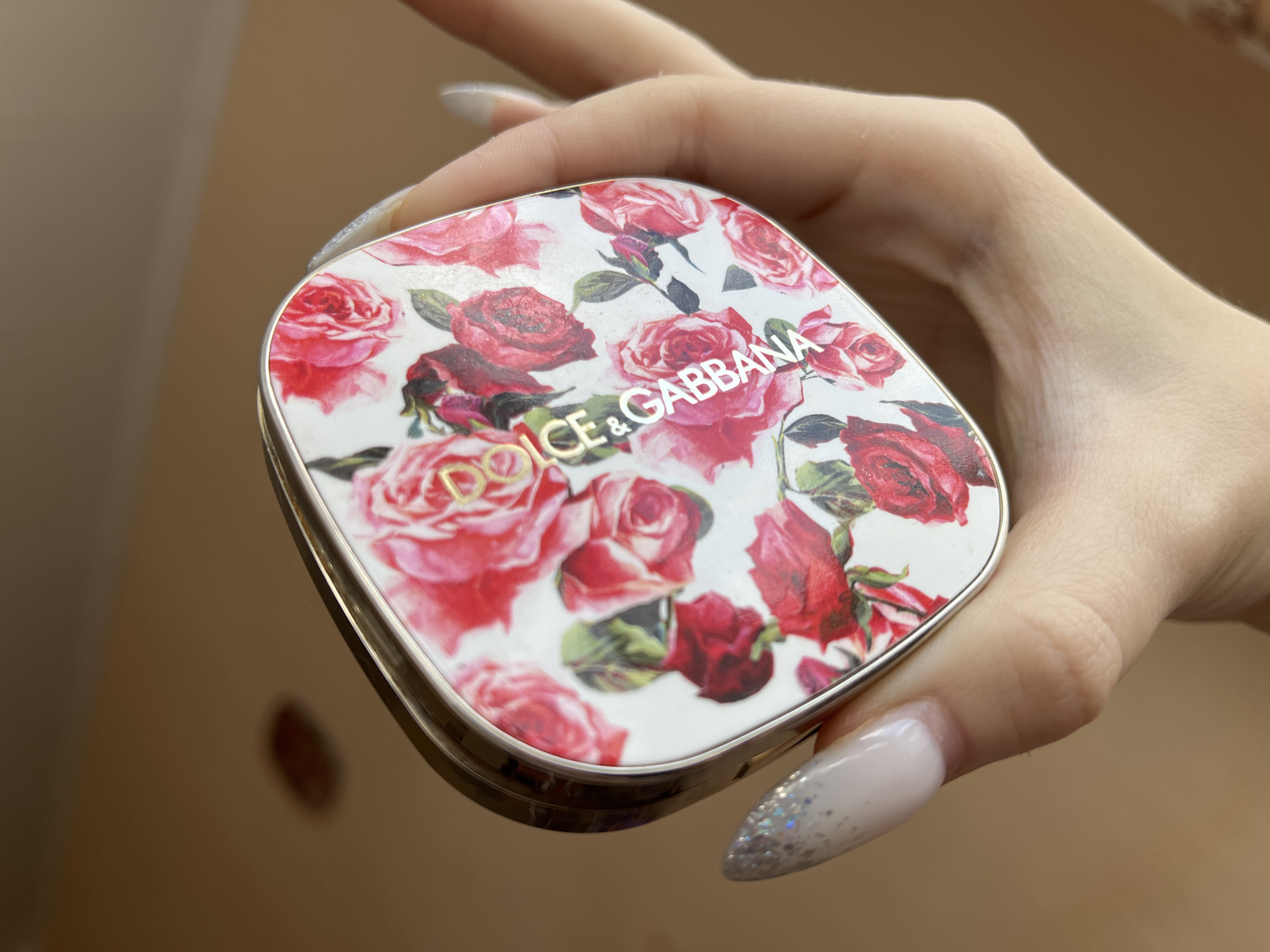 A person's hand is holding a decorative compact with a floral pattern of red and pink roses. The compact has a gold-colored trim and the words "DOLCE & GABBANA" printed in gold on the lid. The person's fingernails are long and have a glittery, pale pink finish. The background is out of focus and appears to be a neutral, warm-toned surface. The image does not contain information about the city of Ivanovo, Russia.