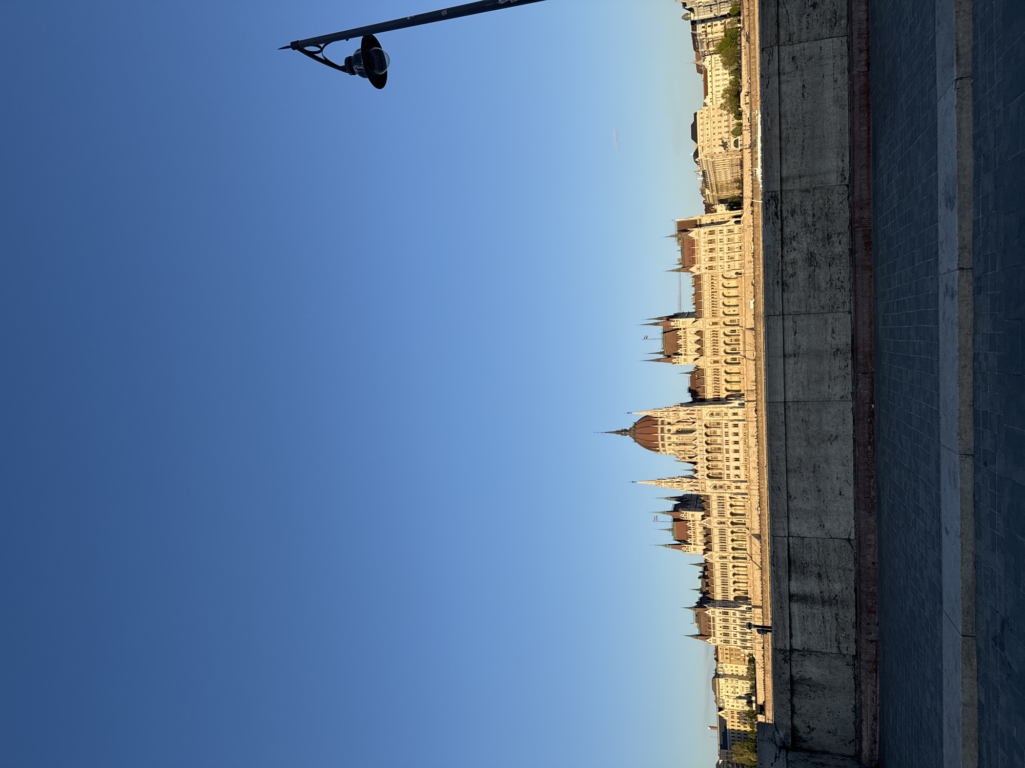 This photograph, originally rotated 90 degrees clockwise, presents a serene view of the Hungarian Parliament Building in Budapest, Hungary. The grand, ornate structure dominates the right side of the frame, showcasing its distinctive Neo-Gothic architecture with numerous pointed spires, a large central dome, and many arched windows. The building's intricate facade is bathed in warm, golden light, indicative of either early morning or late afternoon sunshine.
In the upper left portion of the image, a dark, elegant lamppost extends from the corner, providing a foreground element against the expansive, clear blue sky. Below the Parliament Building, a textured stone embankment runs horizontally, serving as a divide before a paved walkway or path in the lower right foreground. The low sun angle creates subtle shadows, emphasizing the architectural details.
The scene is devoid of people or discernible activity, offering a peaceful and grand perspective of the iconic landmark. The bright blue, cloudless sky suggests fair weather. No visible text is present in the image.