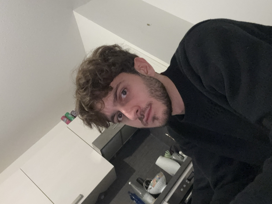A selfie in a kitchen setting features a man with curly brown hair and a light beard. He's wearing a dark sweater. The angle is tilted, showing white cabinets on the left and a stove with various items nearby, including what appears to be a colander and bottles. Cans are visible on top of the cabinets. The kitchen's color palette is mainly white and gray.