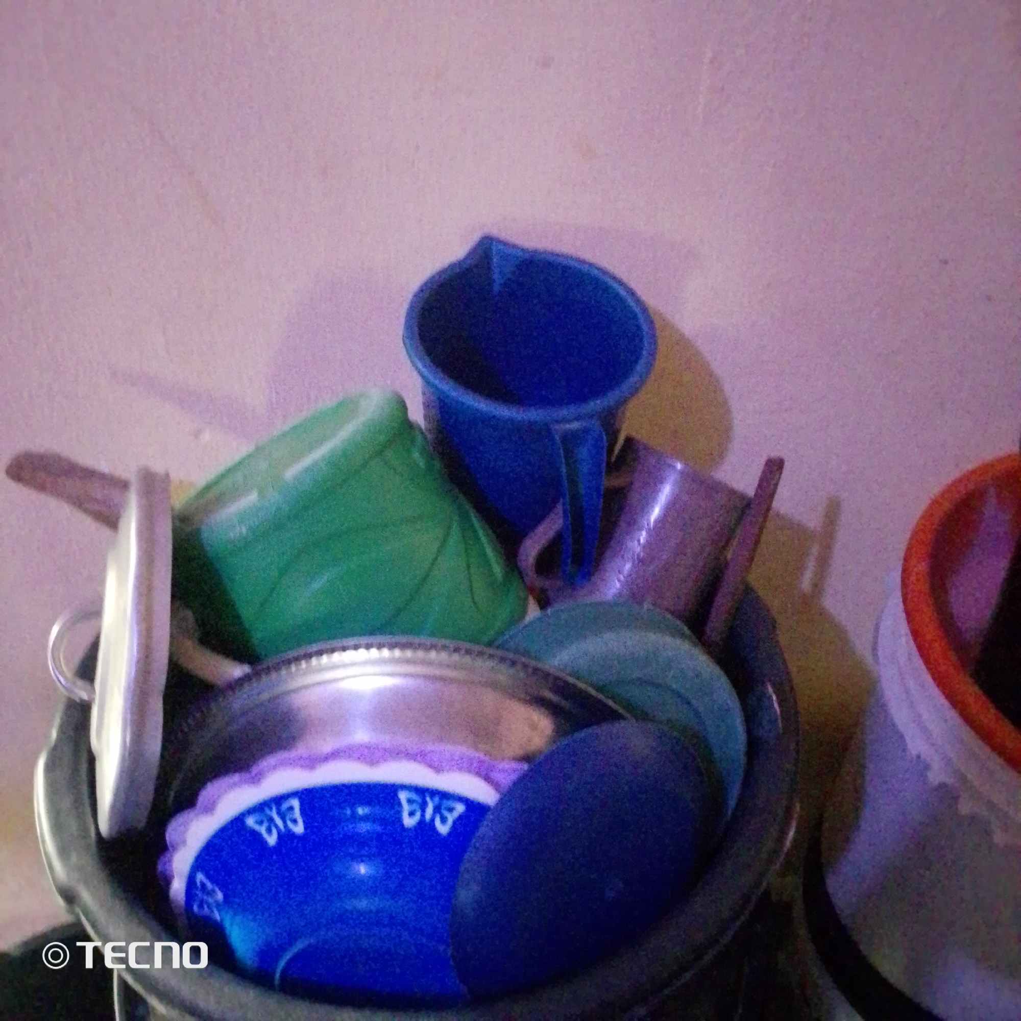 The image depicts a collection of plastic containers and cups piled into a larger, dark-colored bucket or basin, suggesting an indoor setting. The items include a blue mug, a green basin, a purple cup, and a blue bowl with white scalloped edges, possibly containing some white substance. A silver-colored bowl or lid rests on top of some of the items. The background is a plain, textured wall, appearing to be a light pink or purple hue. There are no individuals visible in the image. A watermark "TECNO" is present in the bottom left corner. This scene could be from a home or storage area in Jalingo, Nigeria, where such plastic containers are commonly used for various household purposes.