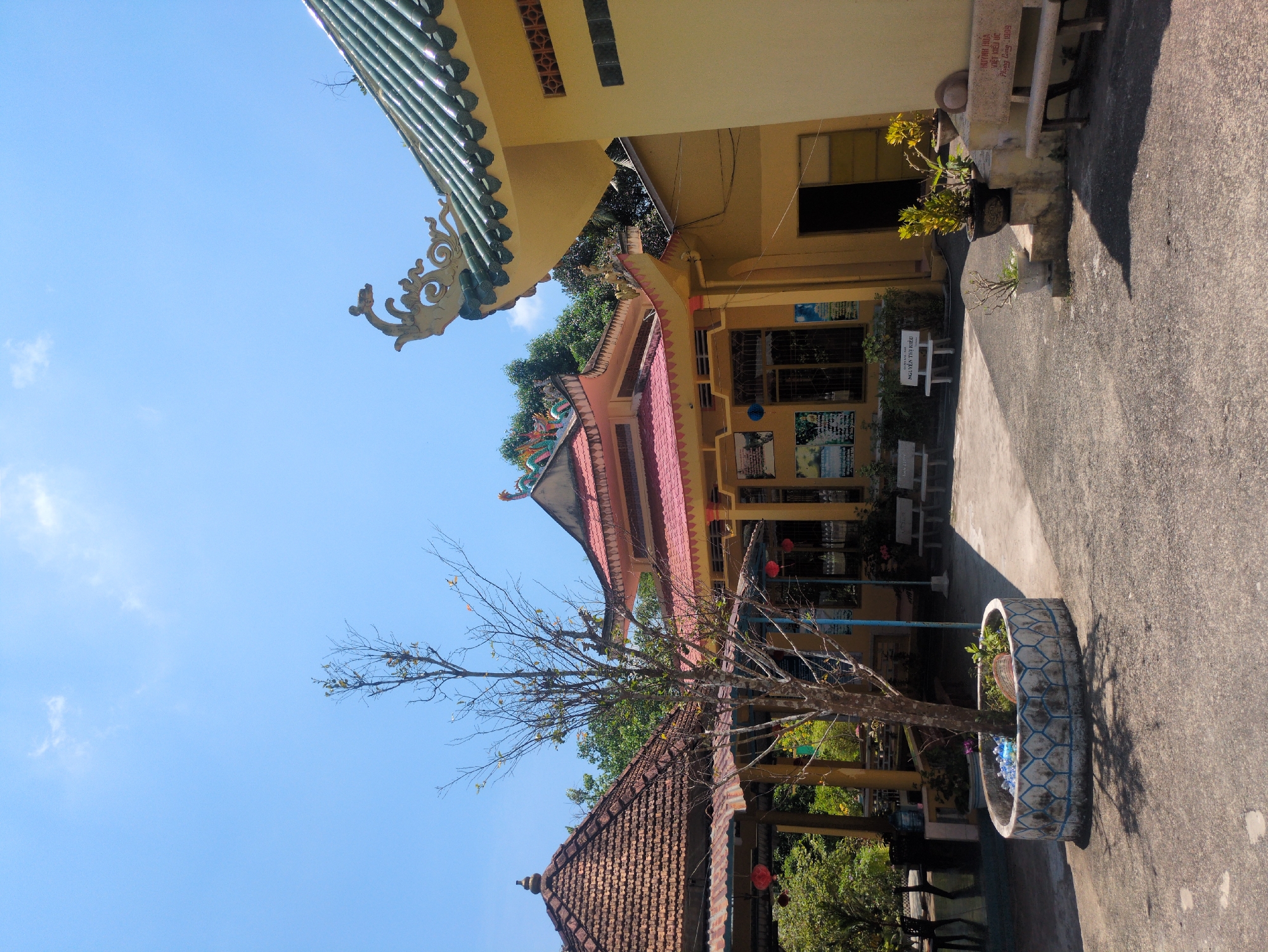 The image depicts a section of a temple complex in Phú Quốc, Vietnam. The architecture features layered roofs with traditional Vietnamese tile work, evident in the green, red, and brown tiled roofs of the various structures. One building is painted yellow and appears to be a newer addition to the complex. A leafless tree stands in a tire repurposed as a planter in the foreground. In the background, temple buildings are visible, including a courtyard area with benches. The sky is blue with scattered clouds.