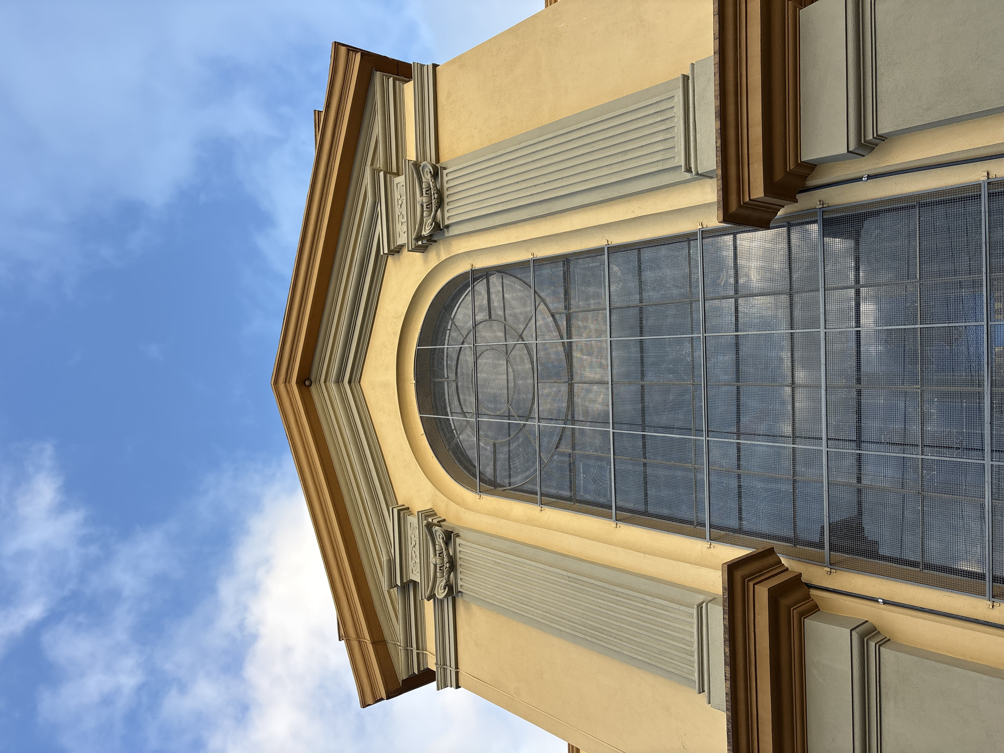 The image shows the exterior of a building with a pale yellow facade and architectural details in grey and brown. A large arched window dominates the center of the building, covered by a wire mesh. The window is segmented into smaller panes by a grid pattern. Above the window, the roofline is visible, with a triangular pediment. Flanking the window are two prominent grey columns with fluted shafts and ornate capitals. The sky above is blue with scattered white clouds. The perspective is from a low angle, looking up at the building. There are no people or any indication of activity. The scene is static and architectural. This image does not provide enough information to determine its location in São Paulo, Brazil.