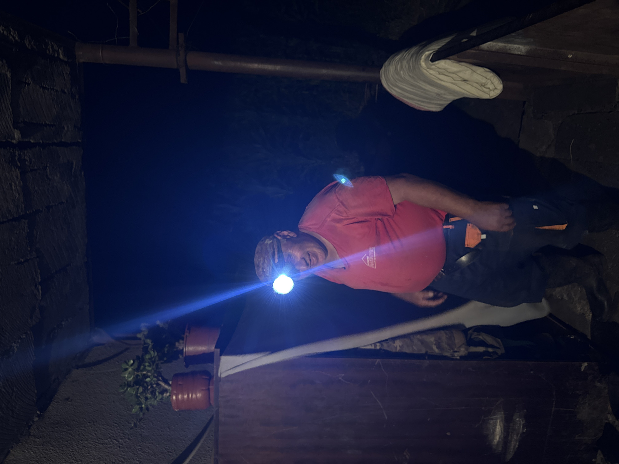 The image depicts a man wearing a red t-shirt and dark pants, with a headlamp illuminating his face, standing in what appears to be an outdoor or semi-outdoor area at night. The headlamp beam is bright white and casts a blueish hue on his face and the surrounding area. He has a smiling expression and is looking slightly to his left.
To his left, there are two potted plants on a shelf, one with lush green foliage in a reddish-brown pot, and another smaller plant behind it in a similar pot. To the far left, a dark, textured surface, possibly a wooden or corrugated metal wall, is visible. A white sheet or fabric hangs down from the ceiling area on the left side of the frame.
In the background, the scene is dark, with hints of foliage or vegetation visible. A vertical support structure, possibly made of metal, is present on the right side of the image. A folded, striped cloth or blanket is draped over a wooden surface on the far right. The ground appears to be uneven, possibly made of stone or dirt. The overall impression is that of a functional, perhaps slightly rustic, environment illuminated by the headlamp. The provided location of Tandzia, Georgia, suggests a rural or semi-rural setting within that region.