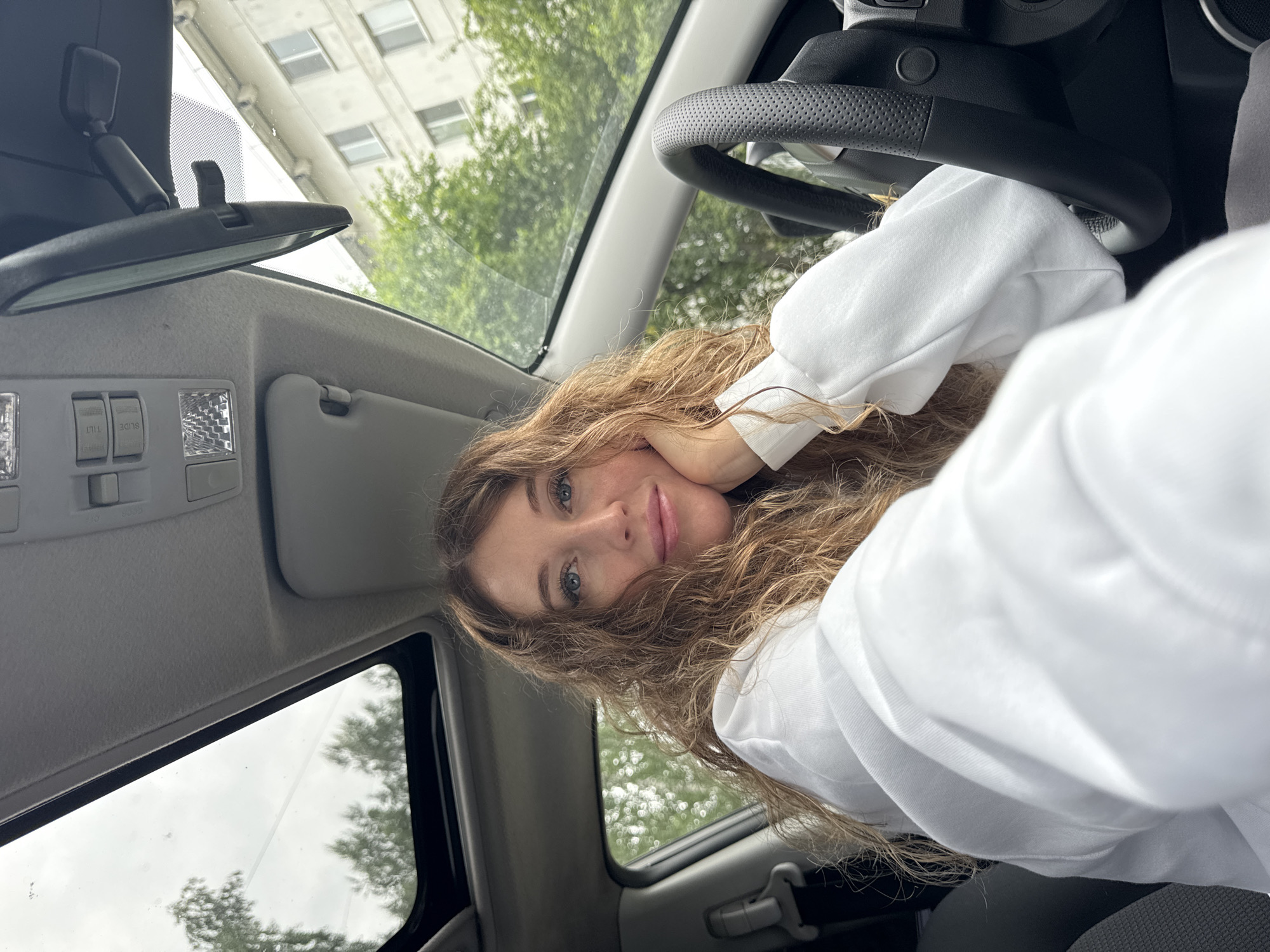 A medium shot shows a woman with long, curly blonde hair and blue eyes sitting in the driver's seat of a car. She is wearing a white hoodie and resting her chin on her left hand. The car's sunroof is open, revealing a cloudy sky and the tops of trees. A building with several windows is visible through the windshield. The steering wheel is on the right side of the car. The scene appears to be in Chaikovsky, Russia, based on the architecture and vegetation.
