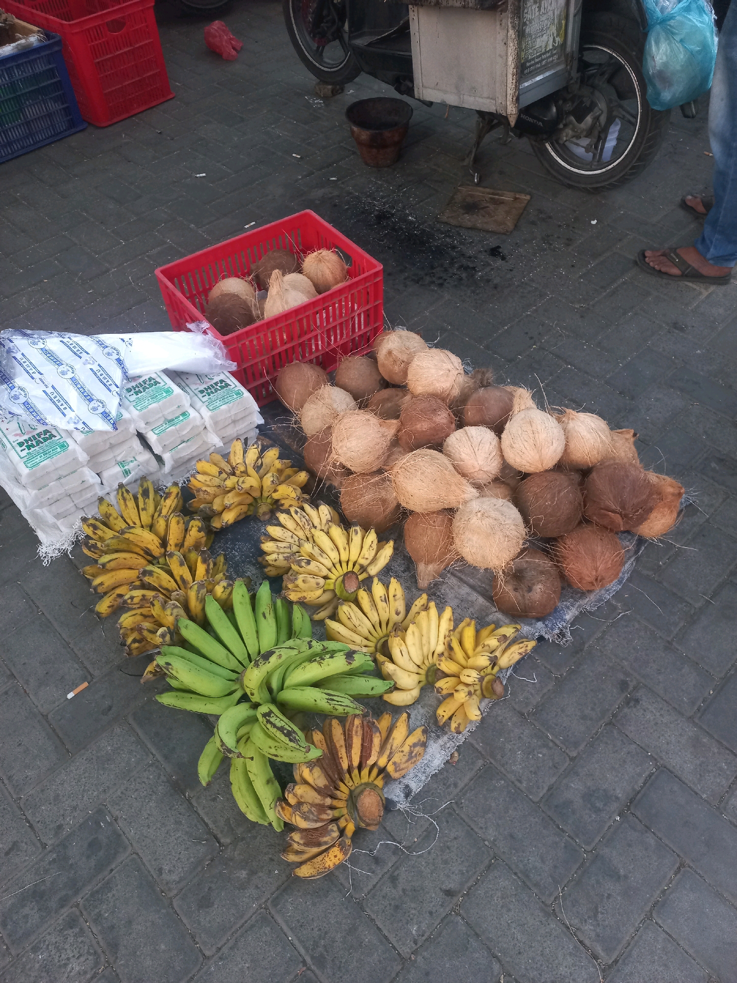 The image depicts a street scene in Baturetno, Indonesia, likely a market area. In the foreground, a variety of fruits are arranged on the ground. There are several bunches of bananas, some appearing ripe and yellow, while others are green. Next to the bananas, a large pile of coconuts is visible. Behind the fruits, several stacks of white packaged goods, possibly food items like sugar or flour, are placed on the ground. In the background, red and blue plastic crates are present. A motorcycle is partially visible, suggesting transportation and possibly a vendor's setup. A person's legs in blue jeans and sandals can be seen on the right side of the frame, indicating human presence. The ground is paved with dark grey interlocking bricks. The overall scene suggests an outdoor market where fresh produce and other goods are being sold.