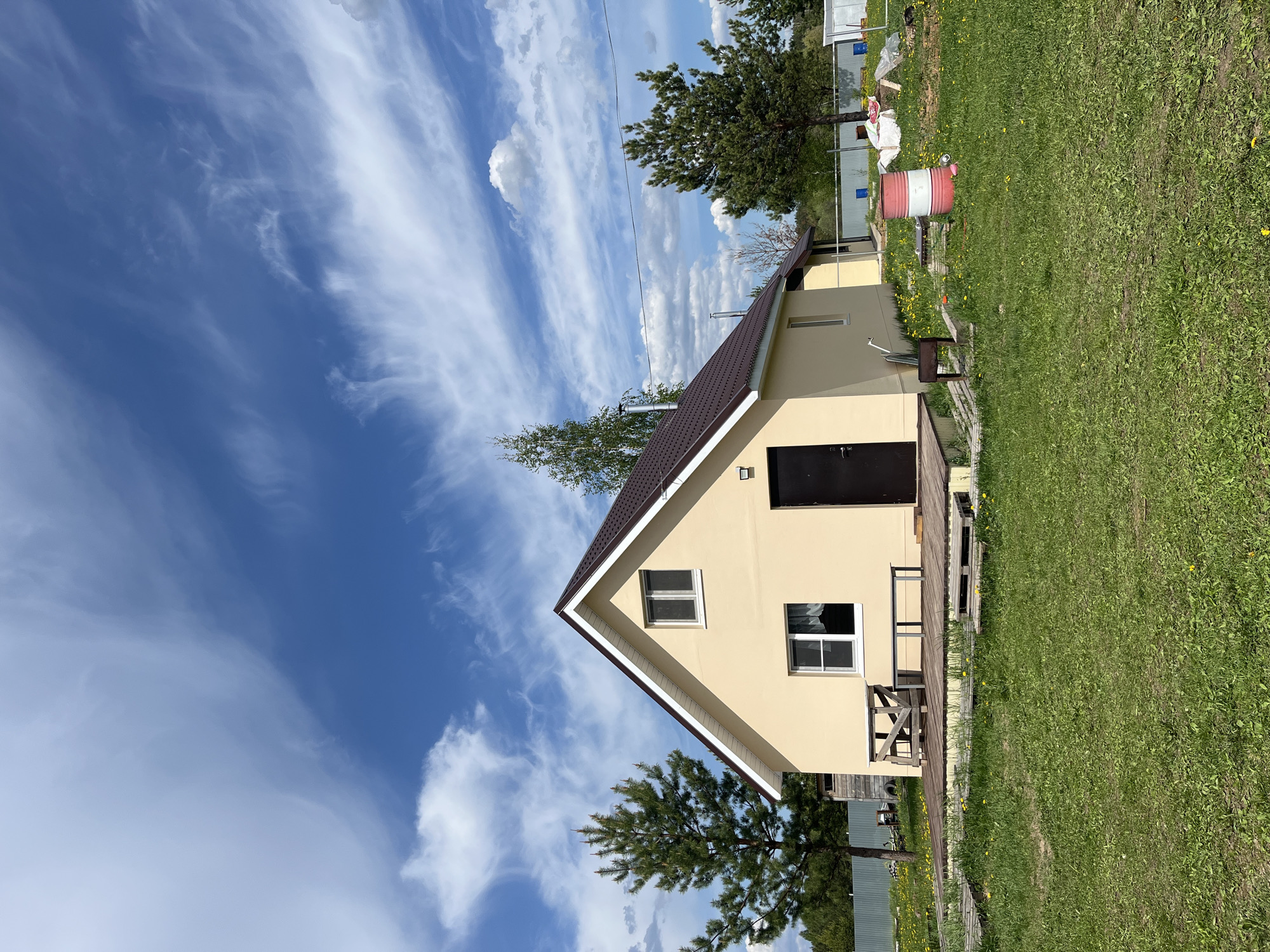 The image shows a single-story house in Гаревая, Russia. The house has a brown roof, light yellow walls, and a dark brown door. There are two windows visible on the front facade. A small wooden deck is attached to the side of the house. The house is surrounded by green grass, and there are trees visible in the background. The sky is blue with white clouds. There is a red and white barrel near the house. A fence is visible further in the background.