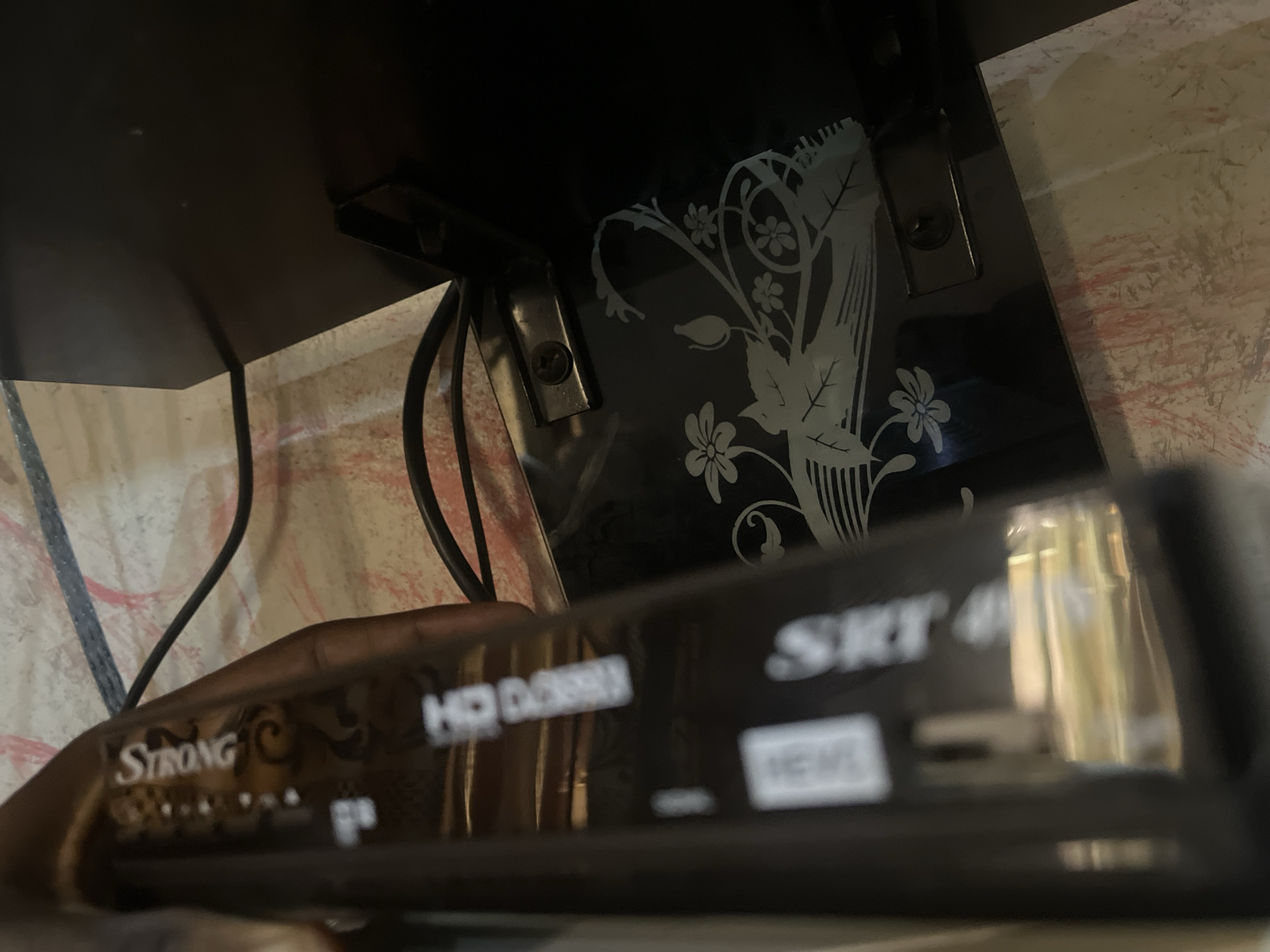 In Jalingo, Nigeria, a close-up shot captures a black satellite receiver labeled "Strong SRT 40" resting on a wooden surface. The receiver has various markings and logos, including "HD DOLBY DIGITAL" and "HDMI." To the left, a hand is partially visible, holding the device. Behind the receiver, a black decorative pattern of flowers and leaves is etched onto the surface of the wooden shelf or cabinet. The background appears to be a wall with a textured, possibly plastered finish, in shades of beige and pink with some red markings. Electrical cords are also visible in the scene, suggesting a connected setup. The lighting is dim, with reflections visible on the glossy surface of the satellite receiver.