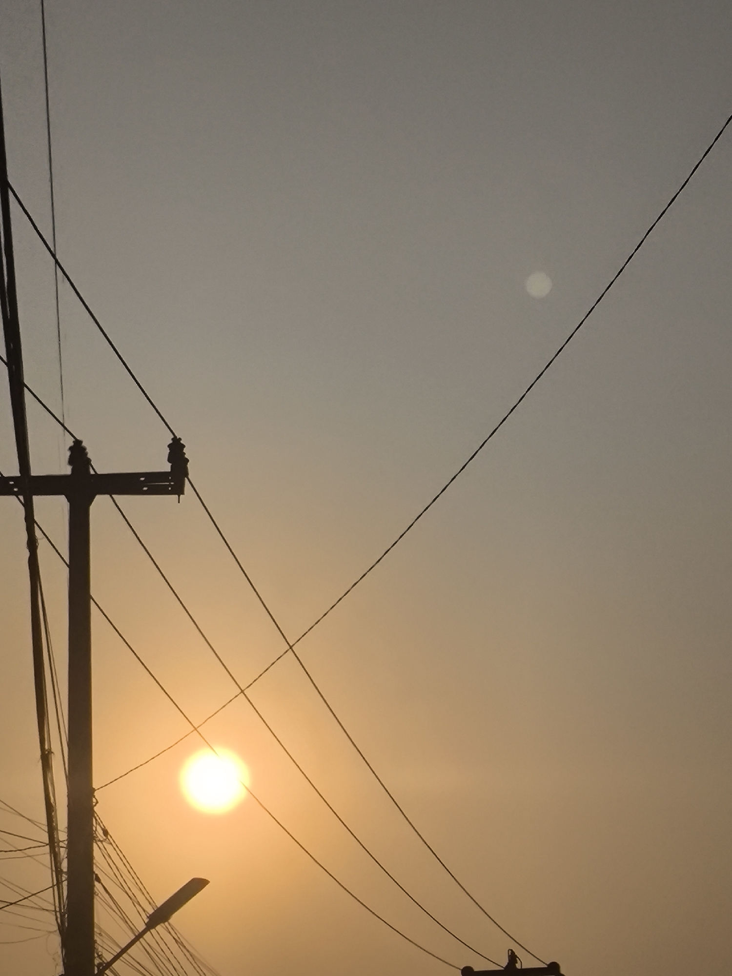 The image depicts a sky scene with the sun setting, casting a warm orange glow. In the foreground, a silhouetted utility pole with numerous wires stretches across the frame. A street lamp is also visible, angled upwards. In the upper right portion of the image, a fainter, possibly moon-like celestial body is visible. The image was taken in Cilegon, Indonesia.