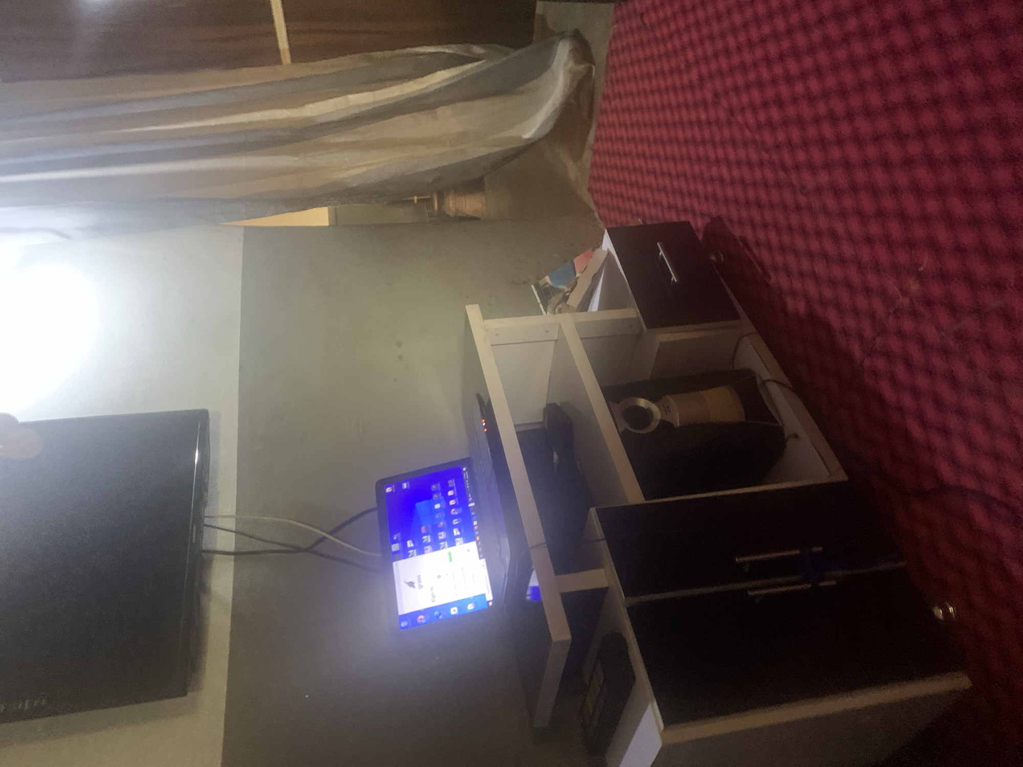 The image depicts a dimly lit room, possibly a bedroom, in Jos, Nigeria. A laptop is open and illuminated on a white shelf unit. The laptop screen displays a login page with a company logo and input fields. The shelf unit also contains various items, including what appears to be a remote control, a small speaker, and some books or magazines. To the left of the shelf, a dark cabinet with silver handles is visible. The floor is covered with a red carpet with a checkered pattern. In the background, a large curtain with vertical stripes hangs near a window, and a television is mounted on the wall above the shelf unit. The overall impression is of a functional, though somewhat cluttered, personal space.