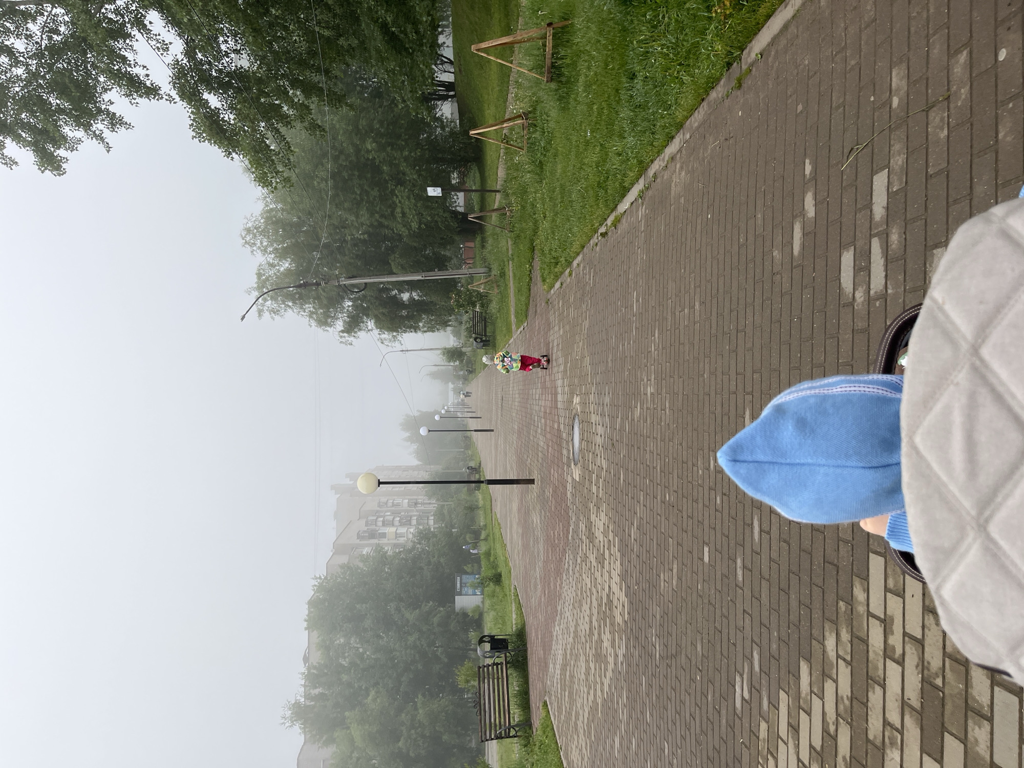 The image captures a misty outdoor scene in Chaikovsky, Russia, taken from a low perspective along a paved walkway. The viewer's point of view includes the feet and lower legs of a person wearing blue pants/shoes and a light-colored, possibly quilted, garment in the foreground on the right.
The path, constructed of brick pavers, extends into the hazy distance, flanked by a strip of grass and trees. Along the grassy verge, there are several park benches, streetlights with spherical lamps, and a few wooden triangular structures, possibly part of an informal play area or landscaping.
Further down the path, a small child is visible in the mid-ground, dressed in a red outfit and a multi-colored hat, seemingly riding a scooter or small bicycle. The background is enveloped in thick fog or mist, obscuring the details of distant buildings, though one large structure is faintly discernible through the haze. A sign with the Russian word "САДОВОД" (Gardener) is visible on a building or kiosk in the middle distance. The weather is very foggy, lending a muted and quiet atmosphere to the daytime scene.