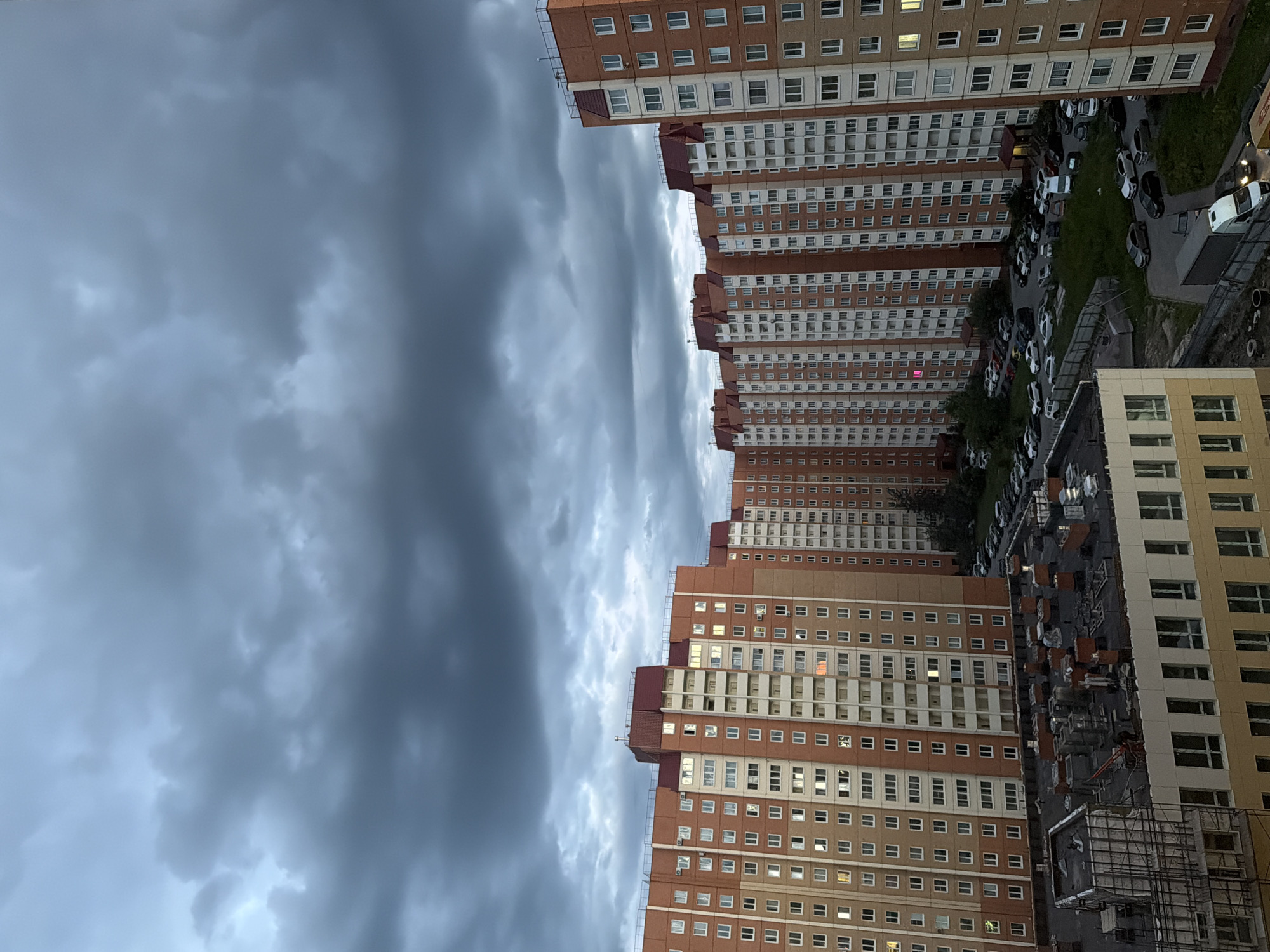 The image shows a dense urban landscape dominated by tall residential buildings with orange and white facades and red pitched roofs. The buildings are arranged in a curve, with many windows visible. The sky above is filled with heavy, dark grey clouds, suggesting an overcast or stormy day. In the foreground, the roof of a lower building is visible, appearing to be under construction with visible scaffolding and equipment. Below the residential buildings, a parking lot is filled with numerous cars of various colors and models. The overall scene depicts a view of a residential district in an urban environment under a dramatic sky.