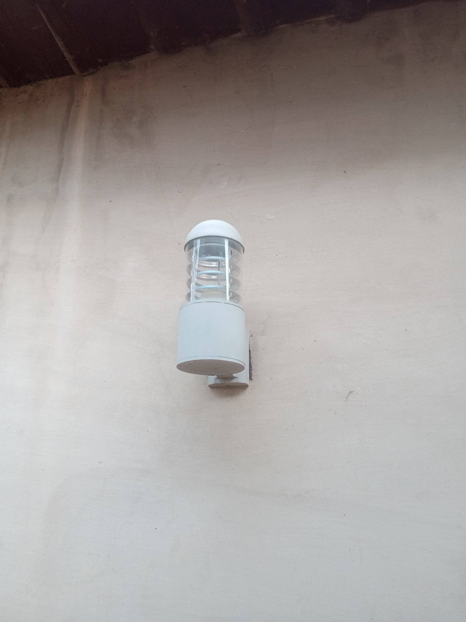 A white exterior light fixture is mounted on a textured, cream-colored wall. The light fixture has a rounded top and a cylindrical base. The glass part of the fixture has a spiral design inside. Above the wall, a dark brown overhang is visible. The scene appears to be an outdoor wall, possibly on a building, in Jalingo, Nigeria.