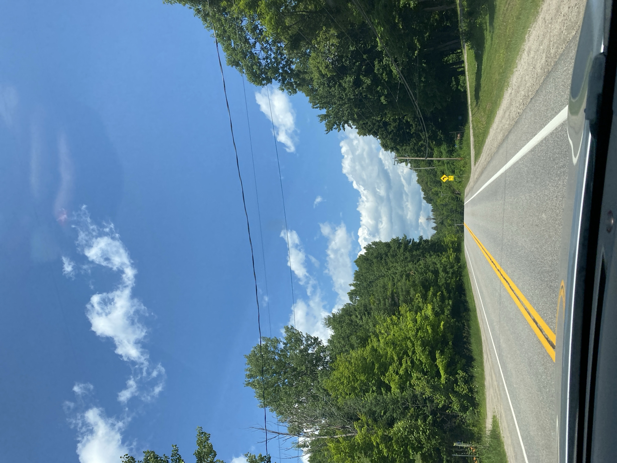 The image depicts a view from inside a vehicle driving on a paved, two-lane road bordered by dense green trees on both sides. The sky is bright blue with scattered white clouds. In the distance, a yellow diamond-shaped sign with the number "75" is visible on the right side of the road, indicating a speed limit. The road features a double yellow line in the center, separating the lanes. The foliage suggests a rural or semi-rural setting, likely during the daytime. The specific location in the city of Lac-Simon, Canada is not ascertainable from the visual information provided.