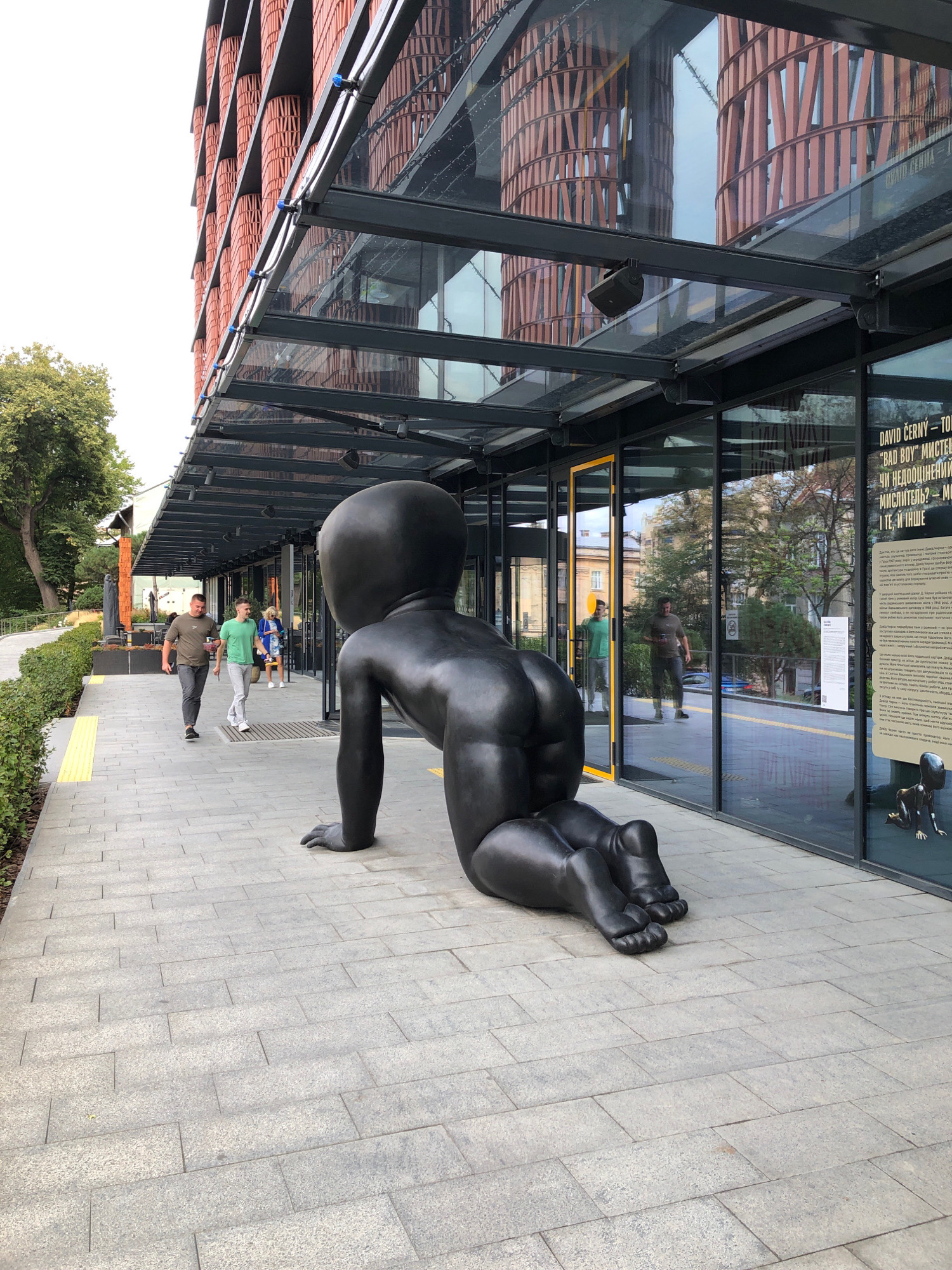 A large, black sculpture of a crawling baby is positioned on a sidewalk in front of a modern building with large glass windows. The baby sculpture faces away from the camera, with its back and buttocks prominently visible. The building's facade features a patterned brick design and reflective glass. On the sidewalk, two men are walking away from the sculpture, and another person is visible further down the sidewalk near the entrance of the building. The environment appears to be an urban setting, possibly a public square or walkway in Lviv, Ukraine, given the presence of trees and the architectural style. The ground is paved with rectangular grey tiles. The overall impression is one of an artistic installation in a contemporary urban space.