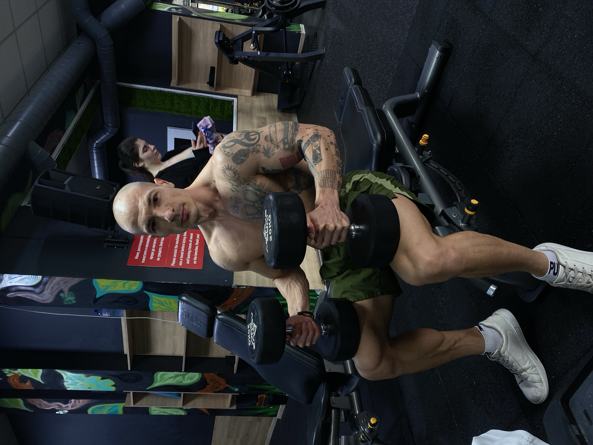 A muscular, bald man with tattoos on his arms is exercising in a gym, lifting two dumbbells. He is wearing green shorts and white sneakers. He is sitting on a weight bench. In the background, another person is sitting near a window. The gym is located in Pleven, Bulgaria, based on the Bulgarian language on a sign visible in the image.