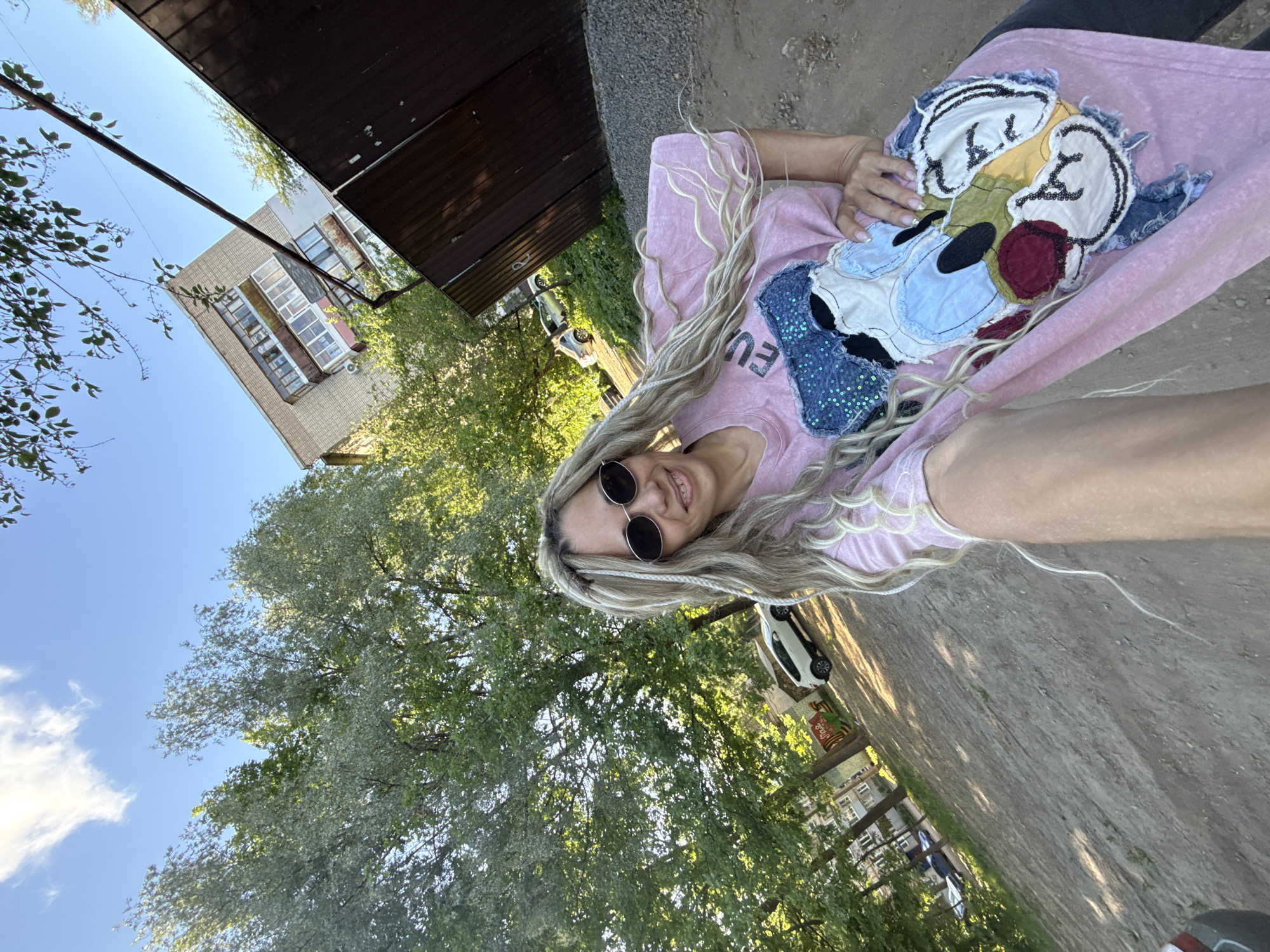 The image shows a woman with long, light-colored hair, wearing sunglasses, a pink t-shirt with a cartoon-like design, and sitting outdoors. The background includes a building, trees, a car, and what appears to be a paved area. The location appears to be Rusanovo, Russia.