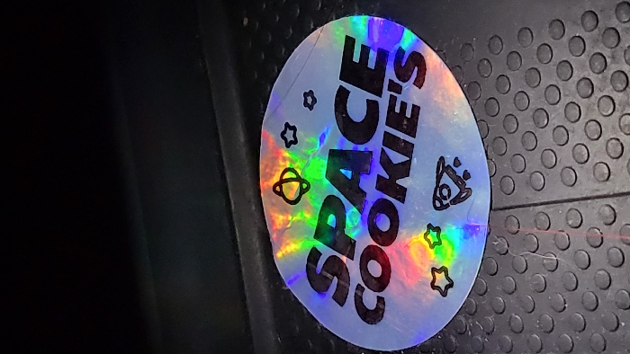 A circular holographic sticker is affixed to a dark, studded, rubberized floor surface in Magaluf, Spain. The sticker displays the black uppercase text "SPACE COOKIE'S," complemented by outline graphics including an atom-like symbol, multiple stars, and a rocket ship with exhaust trails. The holographic material reflects a full spectrum of rainbow colors. Minor creases and surface wear are visible on the sticker. The underlying floor is dark grey or black, characterized by a uniform pattern of raised circular studs, with a faint linear seam present to the right of the sticker. The upper portion of the image is indistinct and dark, indicating a low-light environment.