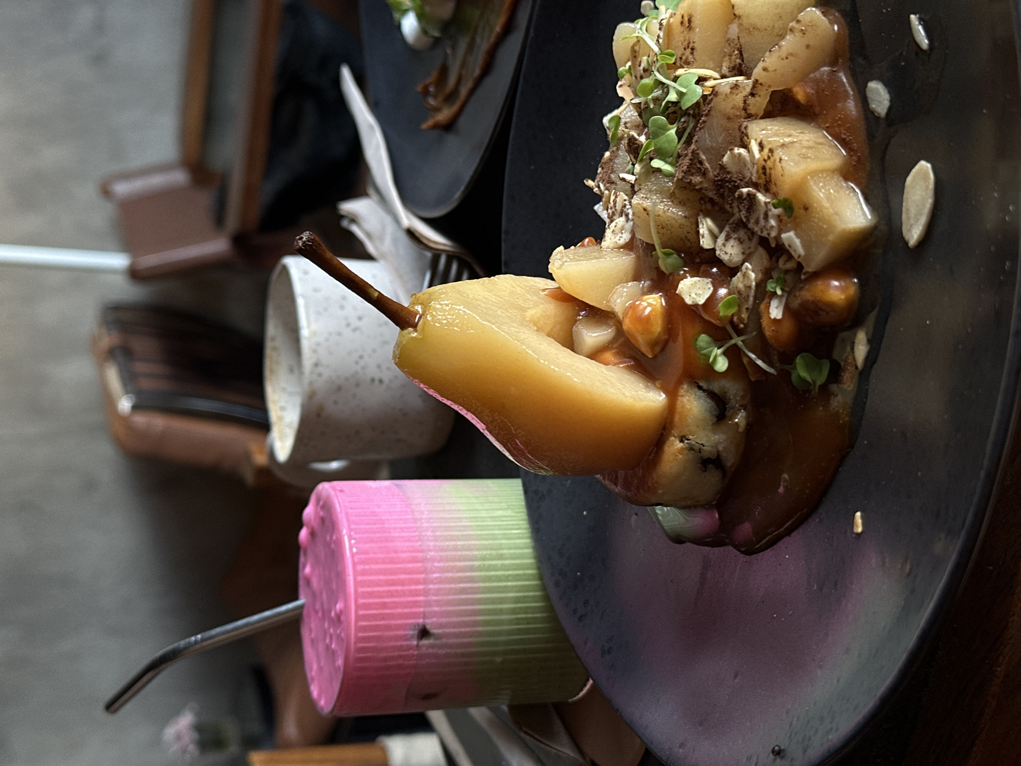 A close-up, overhead shot captures a dessert of poached pears in a dark sauce, garnished with sprouts, slivered almonds, and cocoa powder, served on a black plate. A portion of another black plate with a small dessert is visible in the background. To the left, a white speckled mug with a wooden skewer in it, and a pink and green striped cylindrical object with a metal straw protruding from it. The setting appears to be indoors, possibly a cafe or restaurant in North Kuta, Indonesia, with hints of wooden furniture and a blurred background. The lighting suggests daytime, perhaps late afternoon. The focus is on the food, with no people or prominent emotional cues visible. There is no visible text in the image.