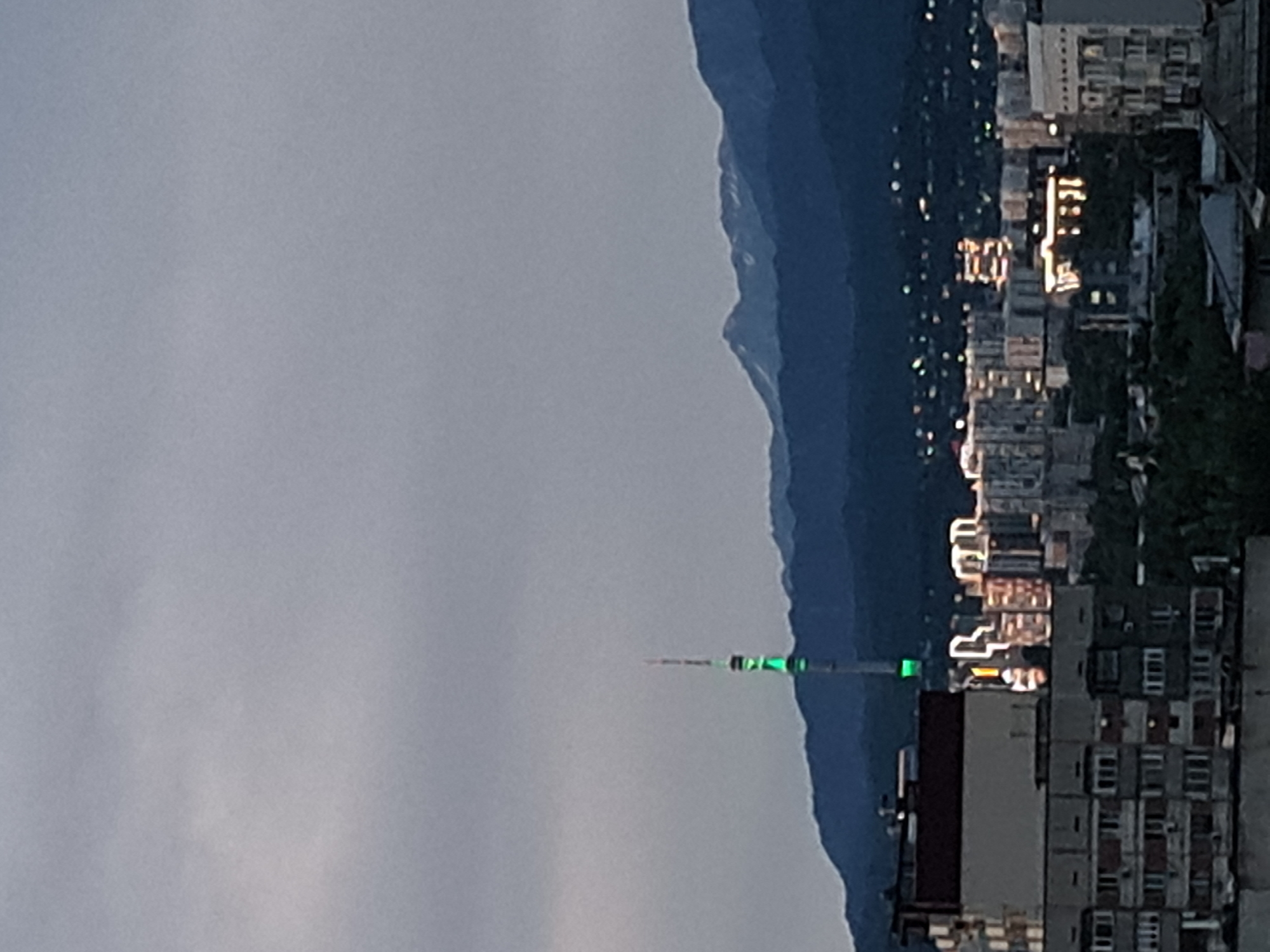 The image shows a cityscape with a prominent television tower in the background, likely the Almaty Television Tower, illuminated with green lights. The tower stands against a backdrop of dark, layered mountains under a cloudy sky. In the foreground, there are several multi-story buildings, some of which appear to be residential blocks with numerous windows. Some buildings in the mid-ground are illuminated with warm lights, indicating a city at dusk or night. The overall scene depicts an urban environment with a natural mountain range as a backdrop, characteristic of Almaty, Kazakhstan.