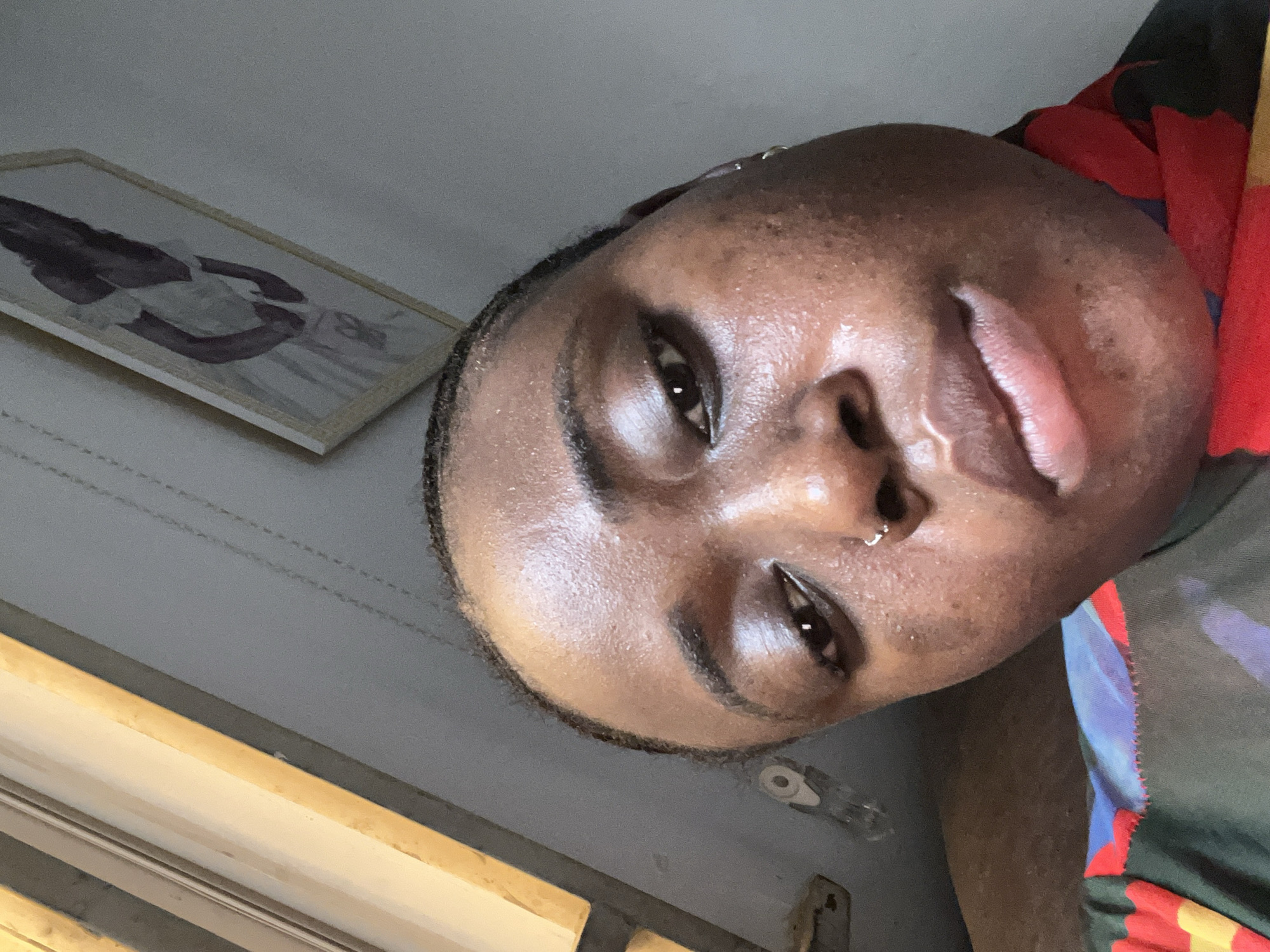 The image shows a close-up of a person's face. The individual has brown skin, dark eyes, and a nose piercing. A framed picture hangs on the wall, and a partial view of a window or a door is visible. The person appears to be wearing a multi-colored shirt. The environment suggests an interior space. There is no information given that this image was taken in Abuja, Nigeria.