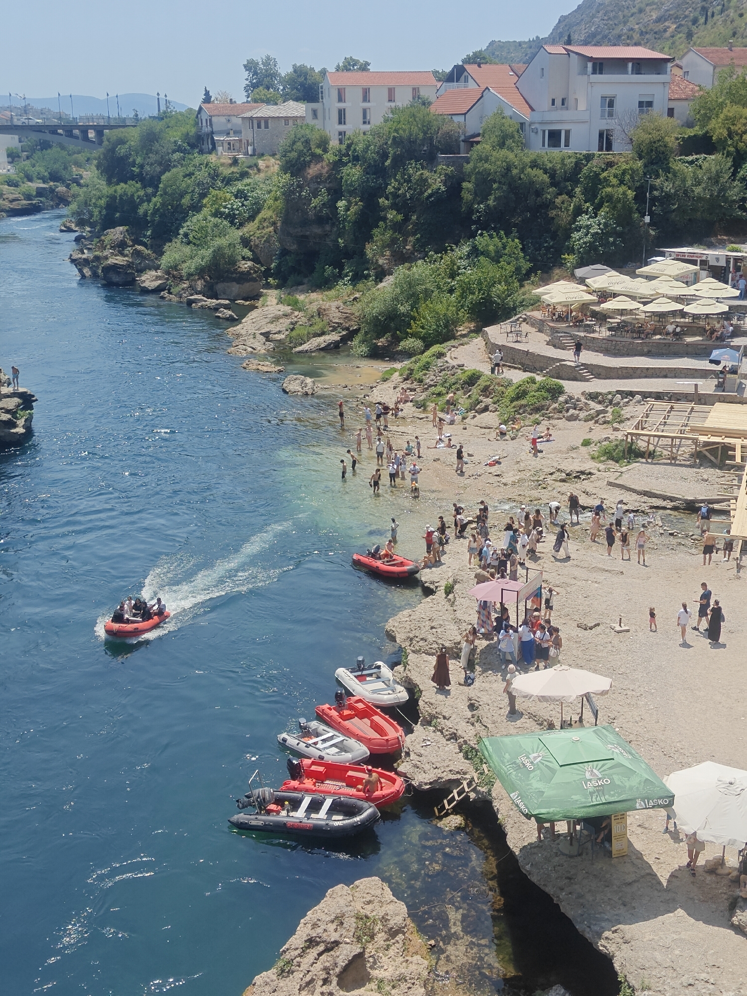 This is an aerial view of a rocky riverbank and the blue water of the Neretva River in Mostar, Bosnia and Herzegovina. On the river, there are several inflatable boats, one of which is moving and leaving a wake, carrying several passengers. Along the riverbank, numerous people are gathered, some wading in the shallow water, others sunbathing or socializing. There are also umbrellas and seating areas set up, suggesting a recreational or dining area. In the background, buildings with red-tiled roofs and lush green trees line the elevated terrain, with a bridge visible in the distance. The overall scene depicts a sunny day with many people enjoying outdoor activities by the river.