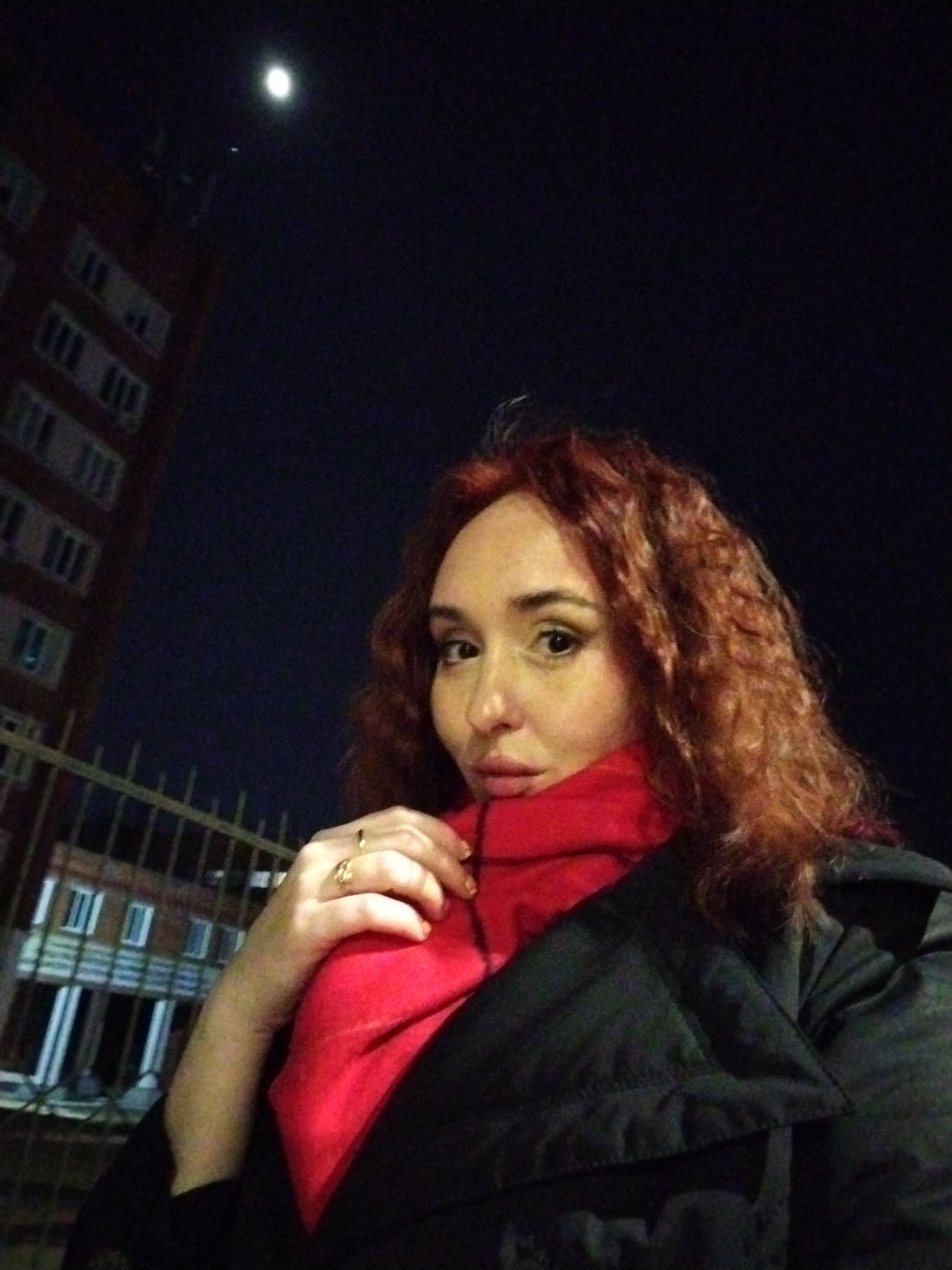 The image depicts a night scene in Izhevsk, Russia, featuring an individual taking a selfie outdoors.
A person with red, curly hair and fair skin is prominently featured in the foreground, occupying the right half of the frame. They are wearing a dark, possibly black, quilted jacket or coat and a bright red scarf wrapped around their neck. The individual's left hand, adorned with at least two gold-colored rings on the ring and middle fingers, is positioned against the red scarf on their chest. Their head is tilted slightly, and their eyes are looking directly towards the camera. One eye is clearly visible, with visible eyeliner.
In the background, a dark, multi-story brick building with multiple rectangular windows is visible on the left side of the frame. Some windows exhibit faint internal illumination. A dark metal fence, possibly a railing or barrier, is present between the individual and the building. The sky is dark, indicating nighttime, with a prominent bright moon visible in the upper left quadrant. A very faint, small light source is also visible below the moon.