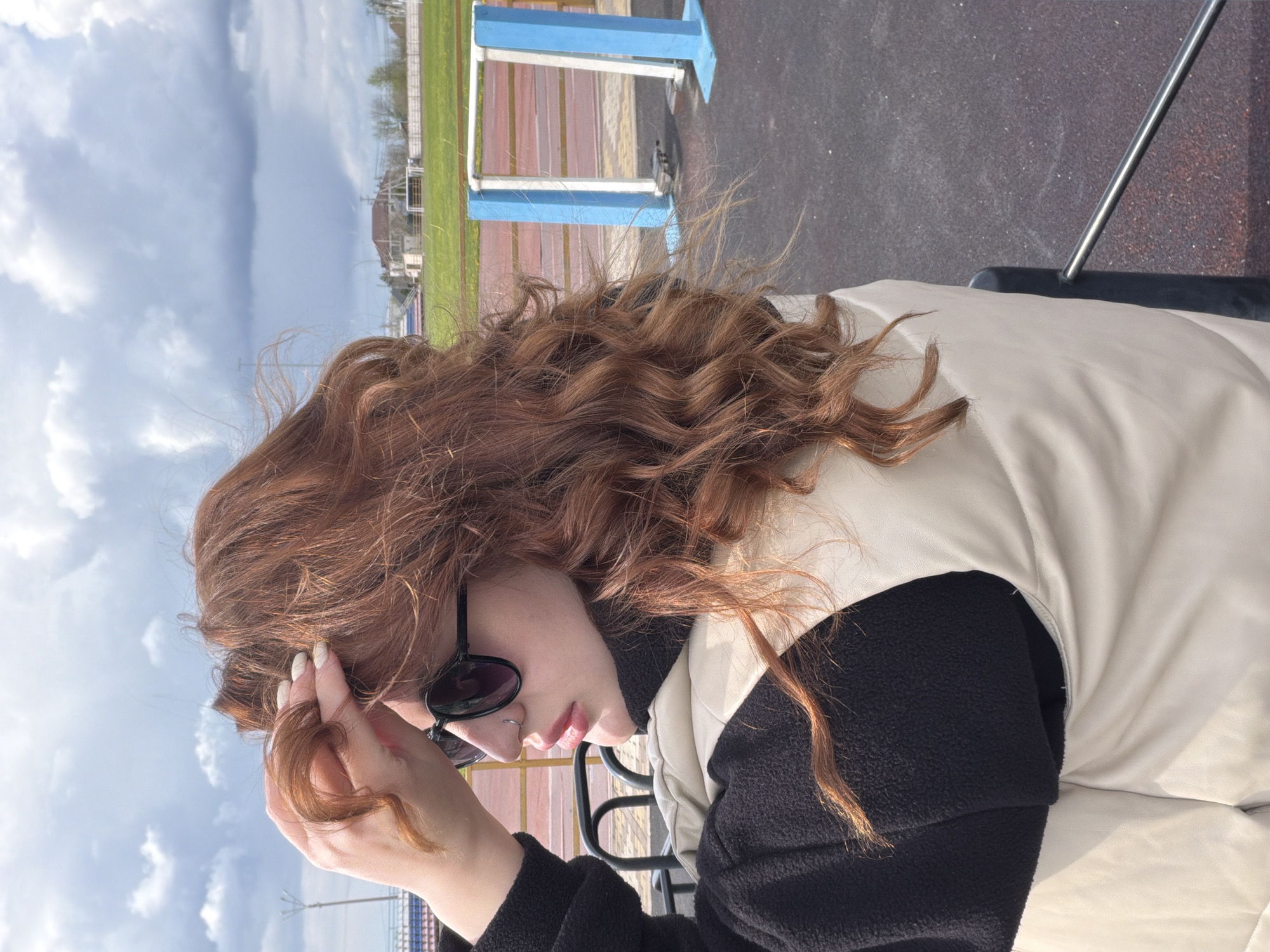 A person with long, curly, auburn hair is sitting outdoors. They are wearing sunglasses, a black top, and a cream-colored vest. The person is touching their hair with their left hand. The background includes a running track, a green field, and a cloudy sky. The location is Novopokrovskaya, Russia.