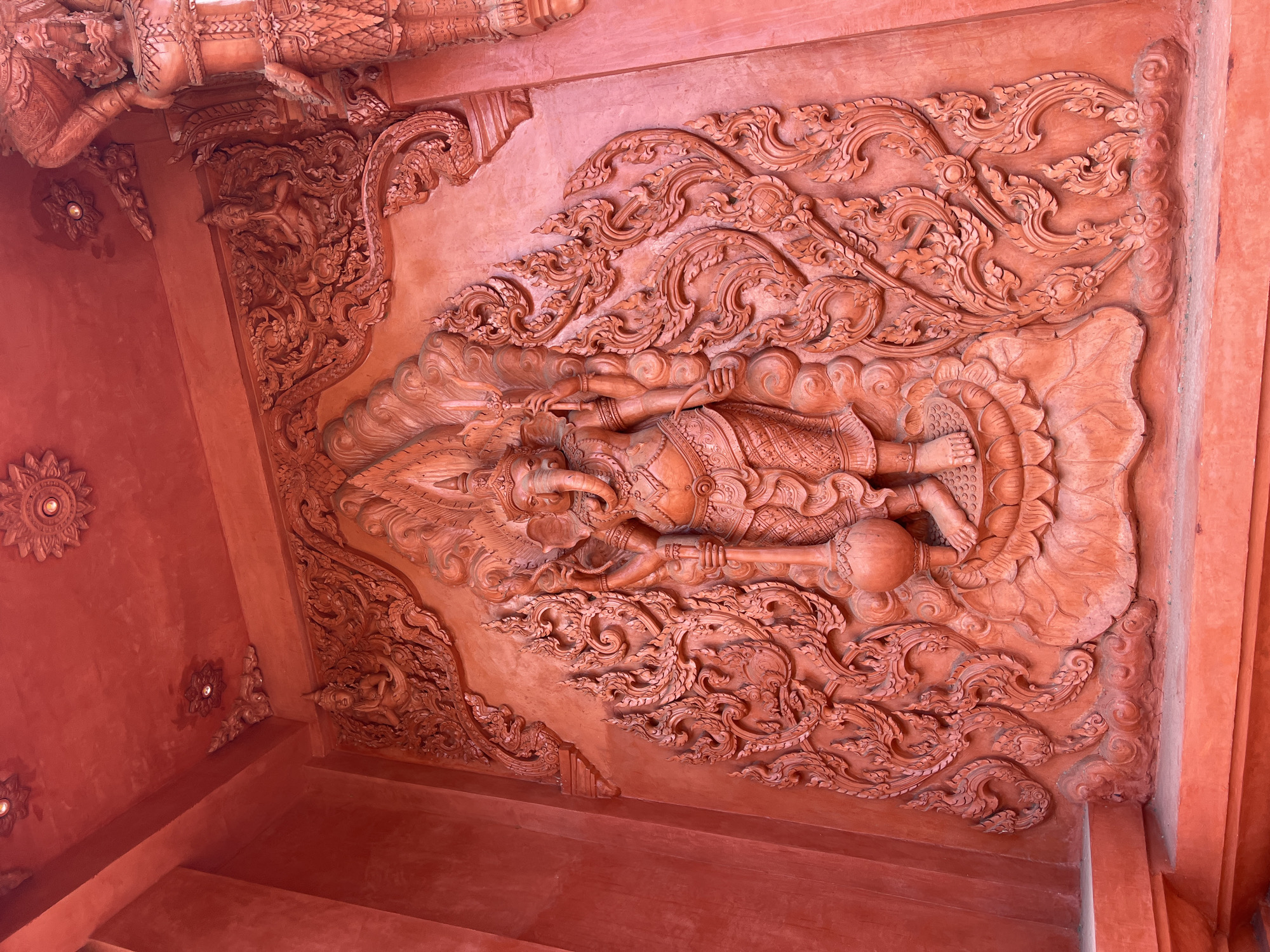 The image shows a close-up of a large, ornate relief carving of the Hindu deity Ganesha, rendered in a vibrant red hue. The carving is set against a textured red wall, likely part of a building in Baan Hua Thanon, Thailand. Ganesha is depicted in a standing pose, adorned with intricate jewelry and clothing, holding a mace-like object in his left hand and a trident-like object in his right. The carving is surrounded by detailed scrollwork and floral motifs, characteristic of Thai temple art. Above and to the right of the Ganesha carving, parts of other large figures, possibly mythological beings, are visible, also in red relief. The ceiling above features decorative rosettes and architectural elements, all in the same striking red color. The overall scene suggests a religious or cultural site, possibly a temple or shrine, with a focus on detailed, monochromatic sculptural decoration.