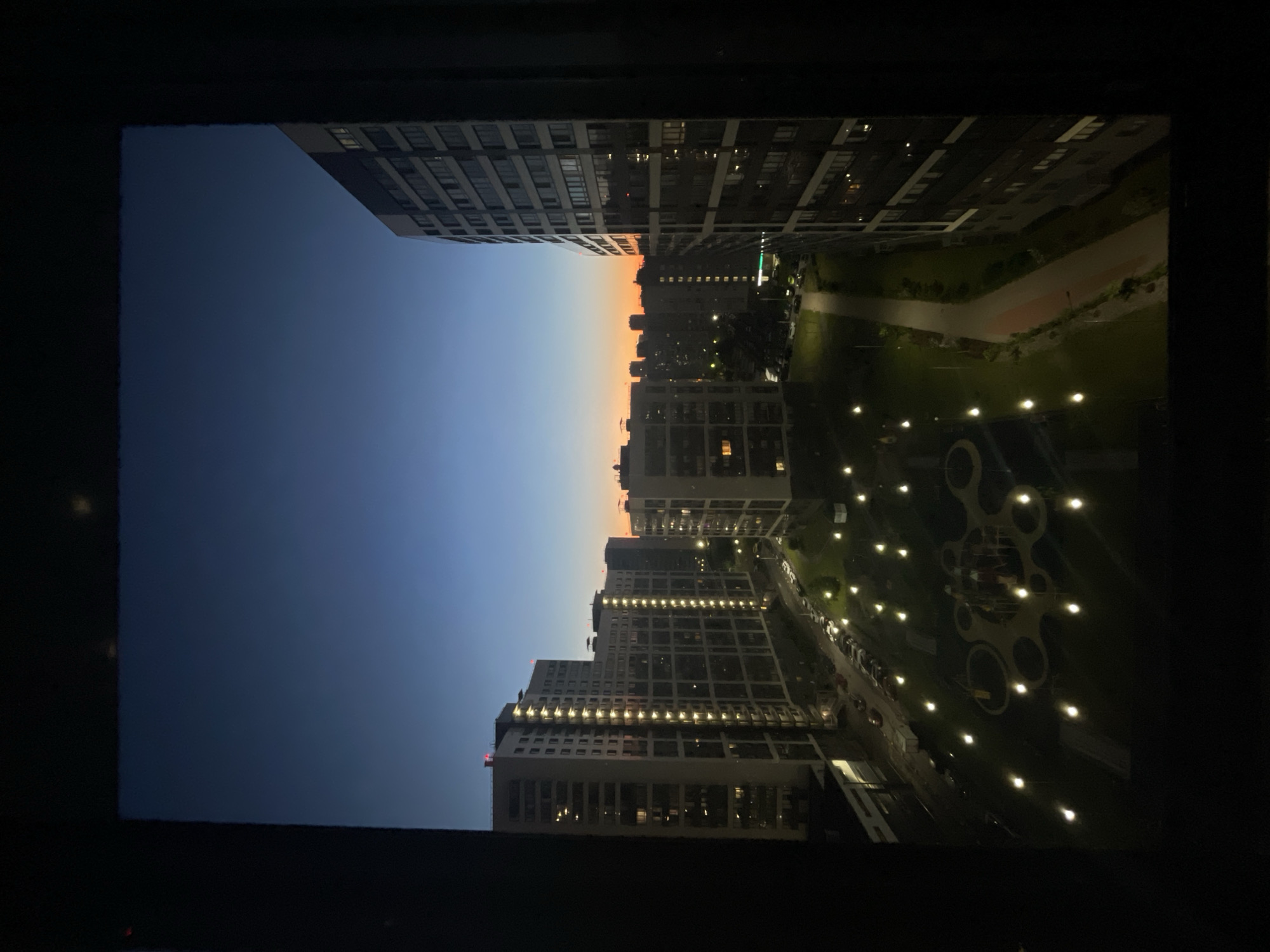 The image depicts a residential cityscape at twilight, as viewed from a window frame. Dominating the scene are several multi-story apartment buildings, characterized by their uniform architectural style and numerous illuminated windows, suggesting the presence of occupants. In the foreground, a dark, irregular frame, likely the window of the vantage point, partially obscures the view.
Below the buildings, a well-lit courtyard area is visible, featuring a playground with colorful equipment and numerous lampposts casting pools of light on the grass and paved pathways. Several cars are parked along a street bordering the courtyard. The sky above transitions from a deep blue at the top to a warm orange and yellow hue near the horizon, indicating either sunrise or sunset. The overall impression is one of urban living in a relatively modern development, with an emphasis on communal outdoor spaces. The location is identified as Sinyavino, Russia.