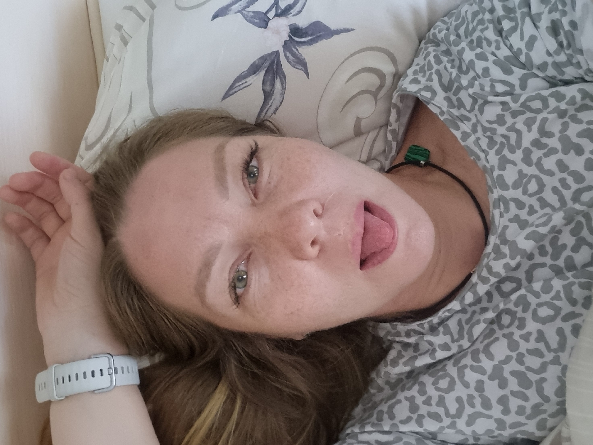 A woman is lying down on a pillow with a floral pattern, with her head turned towards the camera and her tongue sticking out. She is wearing a grey and white patterned shirt. Her right hand is raised near her face, with a white smartwatch on her wrist. She has light brown hair and freckles on her face. A black cord with a green square object is visible around her neck. The location is not identifiable as Chaikovsky, Russia from the image alone.