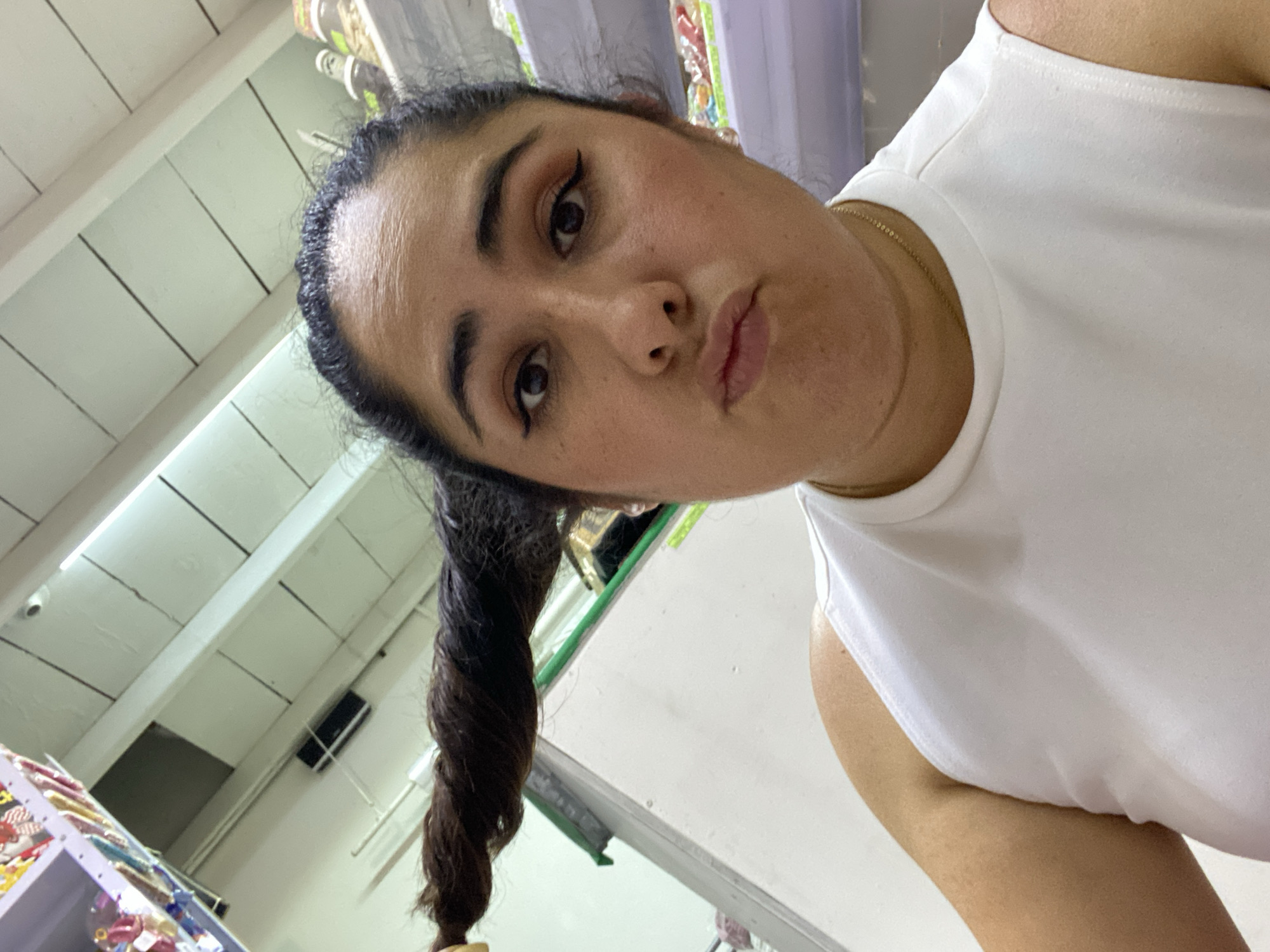 A woman with dark hair twisted into a ponytail is photographed in what appears to be a retail setting, possibly a store. The location is implied to be in San Bernardino Tlaxcalancingo, Mexico, though there's no visible confirmation in the image. She is wearing a white sleeveless top and is looking directly at the camera with a pouting expression. The background shows shelves with merchandise. The lighting in the room is bright.