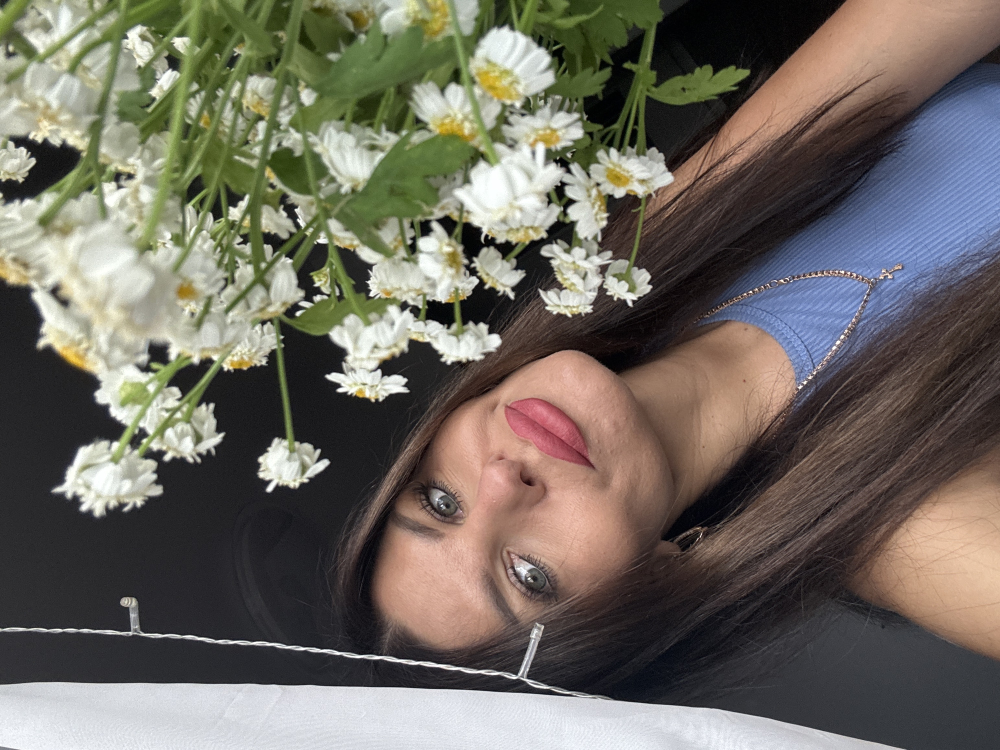 The image shows a close-up of a woman with long, brown hair and blue eyes, wearing a blue top and a gold necklace with a cross pendant. She is looking at the camera with her head tilted. In the foreground, there is a bouquet of white flowers with yellow centers. A white sheer curtain and string lights are visible on the left side of the image. The background is dark and indistinct, suggesting an indoor setting. The image does not contain any information that would allow for a specific location in the city of Chaikovsky, Russia to be identified.