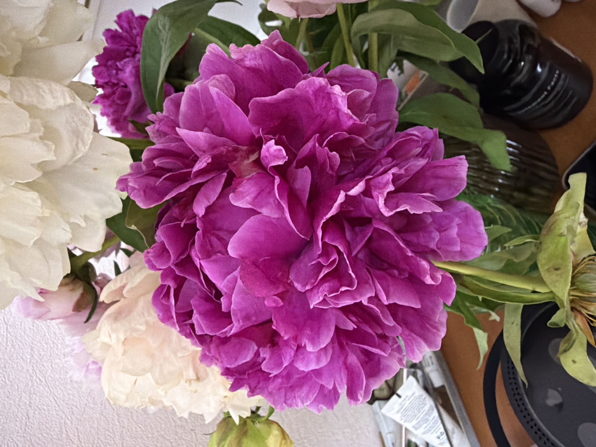 A close-up, slightly overhead shot features a bouquet of peonies, dominated by a large, vibrant magenta bloom in the foreground. Surrounding this central flower are several other peonies in shades of cream and soft pink, as well as lush green leaves and stems. The bouquet is arranged in a clear glass vase, partially visible at the bottom center. The background consists of a textured, off-white wall. The lower portion of the image shows a wooden surface with various objects, including a black circular object with a textured rim, a dark-colored jar with ribbed detailing, and a white tube with black text. The overall lighting is soft and natural. The scene is indoors.