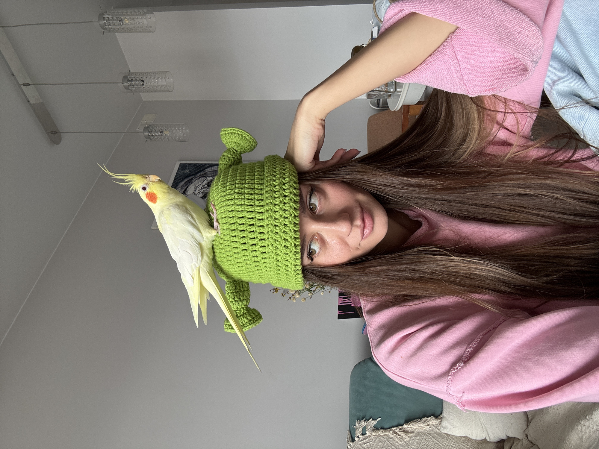An indoor portrait from Krasnoyarsk, Russia, shows a young woman with long, flowing brown hair, captured from a slightly overhead, tilted perspective. She wears a casual bright pink long-sleeved top and a distinctive light green crocheted hat with two rounded, ear-like protrusions, resembling a Shrek-inspired design. Perched comfortably on top of her hat is a yellow cockatiel bird, characterized by its prominent crest and orange cheek patches. The woman gazes directly at the camera with wide, expressive eyes and a soft, engaging smile, conveying a playful or surprised emotion. Her right hand is raised, with fingers lightly touching the side of her head.
The background consists of a plain light gray wall. Several modern ceiling lights with clear, textured cylindrical shades, suspended by thin wires, are visible at the top left of the frame. Towards the right, glimpses of a light blue fabric, a dark green cushion, and a patterned, lighter-colored textile suggest a relaxed domestic setting. A small cluster of dried flowers or foliage can be seen near her hair, behind the hat. The lighting is soft and ambient, indicative of an indoor environment. There is no visible text in the image.