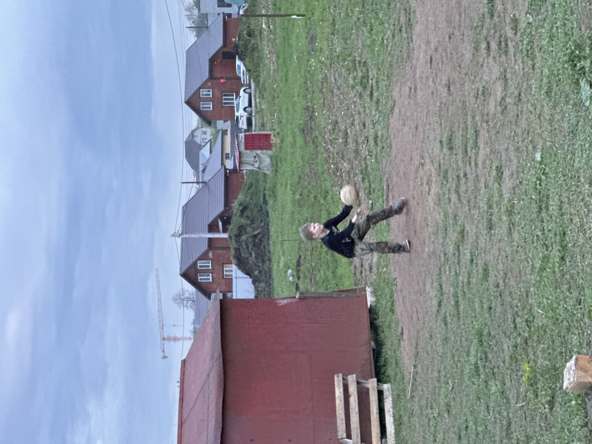 A person is throwing a ball in a grassy area in Ферма, Russia. The person is wearing a dark-colored long-sleeve shirt and camouflage pants. A red building is in the foreground. Row houses are visible in the background. A construction crane is also visible. The sky is overcast.