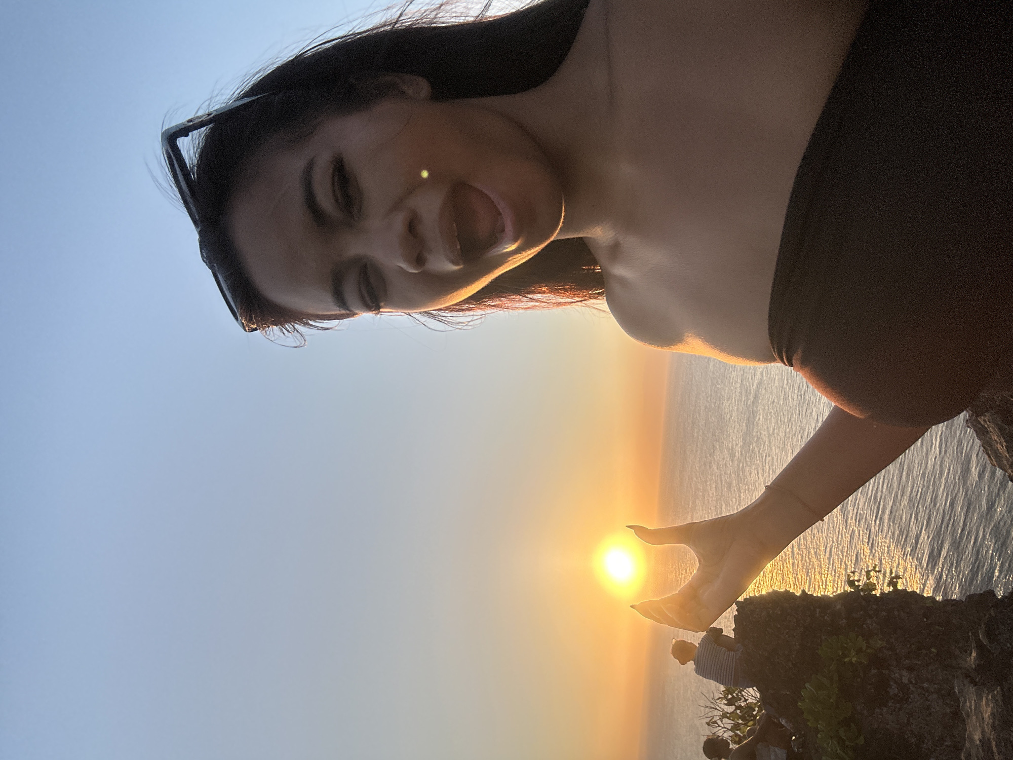 The image captures a young woman taking a selfie on a cliff overlooking the ocean at sunset. She is holding her hand up, cupping the sun as if to hold it, a common pose at Uluwatu Temple in Kuta Selatan, Indonesia. Her mouth is open in a surprised or excited expression, and sunglasses are perched on her head. In the background, two other individuals are seated on the cliff edge, silhouetted against the vibrant orange and blue hues of the sunset. The calm sea stretches to the horizon, reflecting the warm light of the setting sun. The scene is set on a rocky cliff face with some vegetation growing on it.