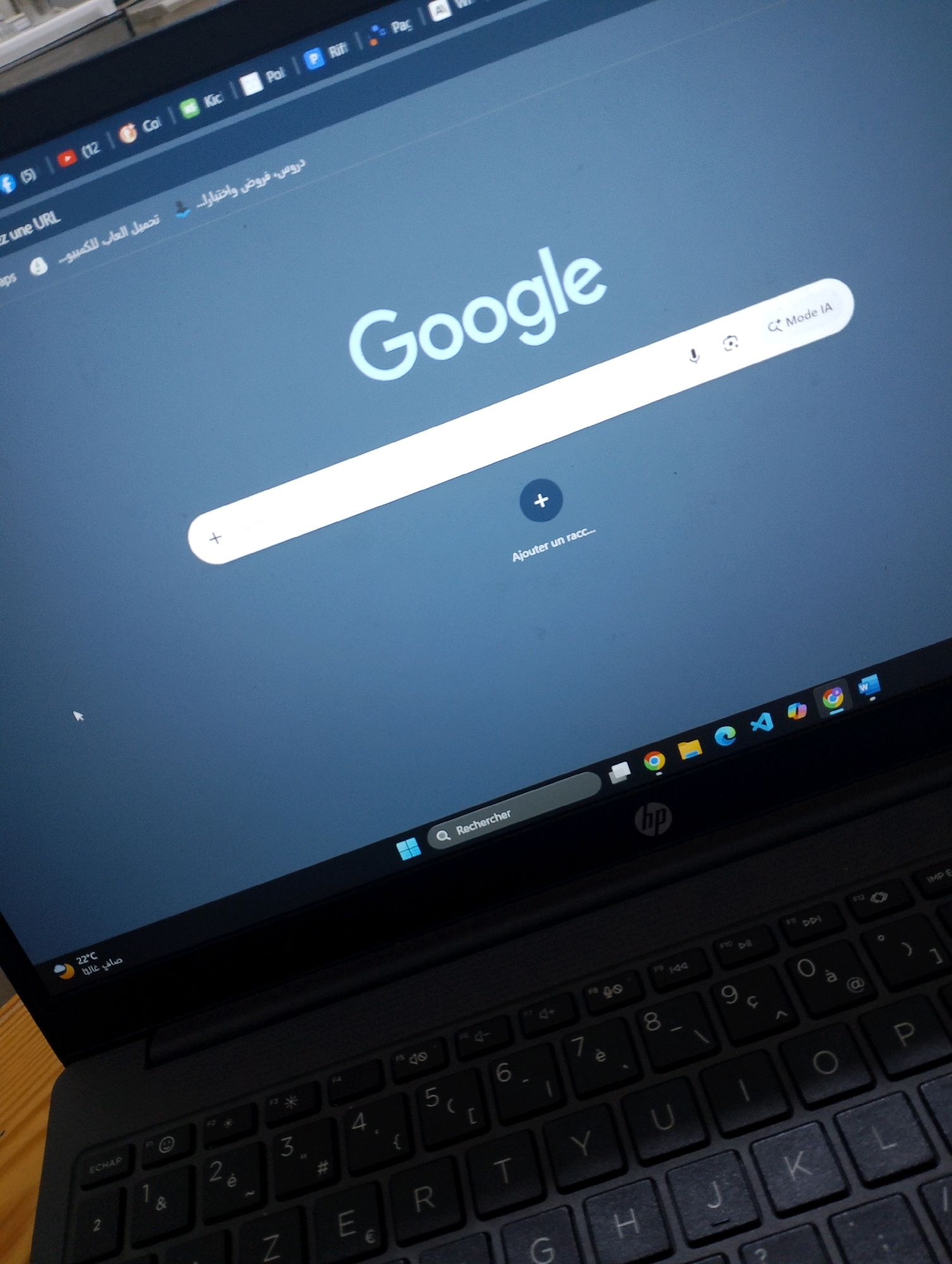 The image displays a close-up view of a laptop screen open to the Google homepage. The laptop is positioned on a wooden surface, suggesting it's in a home or office environment. The screen shows the familiar Google logo and search bar. In the lower part of the screen, a taskbar is visible with various application icons, including a search bar labeled "Rechercher" (Search in French). On the left side of the taskbar, there's a small display showing "22°C" and Arabic text, possibly indicating the local weather and location. The laptop appears to be a standard model, dark in color, with a visible keyboard in the foreground. There are no people in the image, and the action depicted is passive, simply displaying the computer's interface. The overall impression is a snapshot of a digital workspace. The presence of Arabic text and the temperature display suggest the location is likely within Algeria, and potentially in Bordj Menaiel if that context is provided.