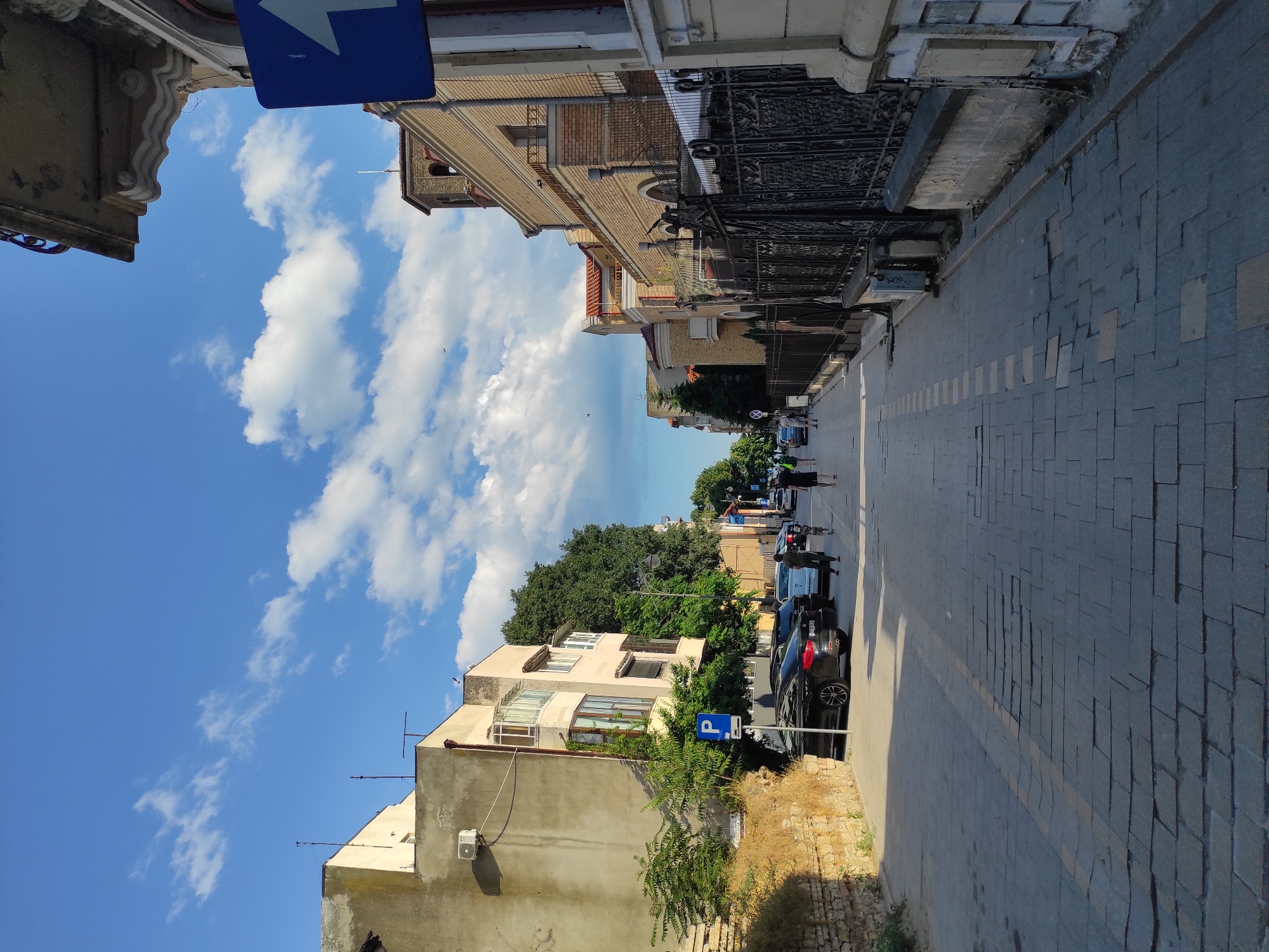 The image depicts a street scene in Constanța, Romania, under a bright blue sky with scattered white clouds. The street is paved with cobblestones and lined with buildings on both sides. On the left, a pale yellow building with several windows and a protruding bay window stands, with a brick wall and some dry vegetation in the foreground. A parking sign with a blue 'P' is visible next to the sidewalk. Several cars are parked along the street, including a dark-colored sedan. On the right side of the street, a series of buildings with yellow brick facades and red-tiled roofs are visible. A decorative black metal fence runs along the front of these buildings. A blue traffic sign with a white arrow pointing upwards is mounted on a pole on the right side of the street, near the foreground. Several pedestrians are walking along the sidewalk. The overall impression is that of a sunny day in a European city.