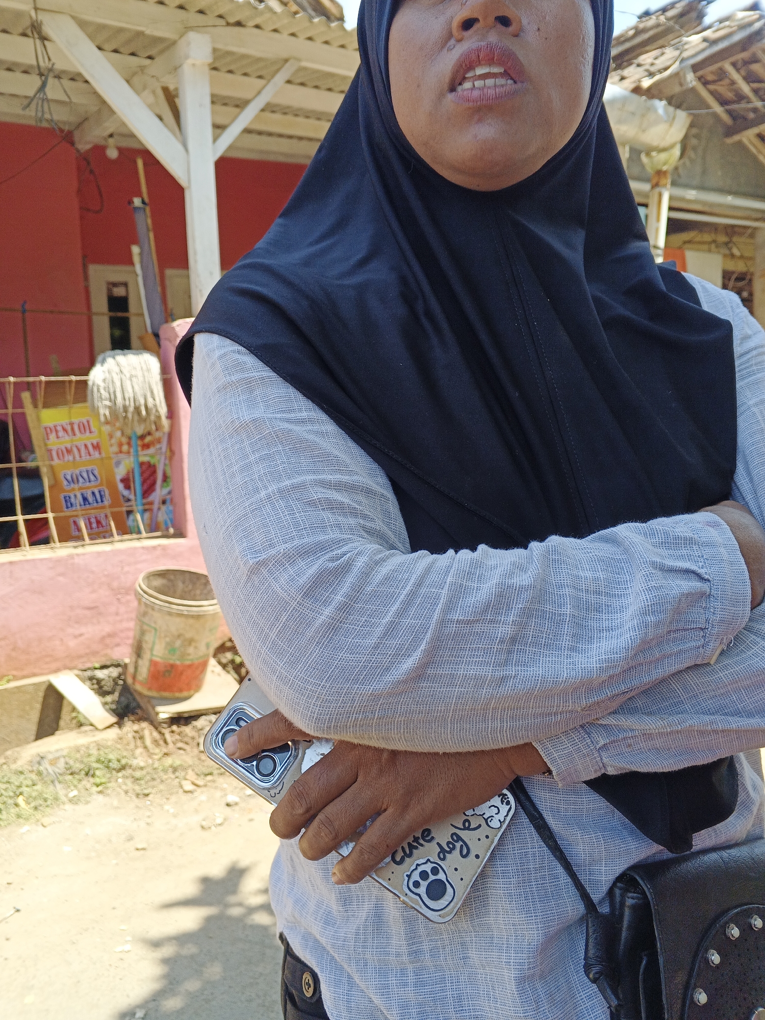 An Indonesian woman wearing a black hijab and a light blue, textured long-sleeved shirt stands with her arms crossed. She is holding a smartphone with a decorative case featuring a drawing of a dog and the text "cute dog". A black leather bag with silver studs is visible on her right side. In the background, a pink building with white structural beams is partially visible, along with a sign in Indonesian advertising food items such as "PENTOL", "TOMYAM", "SOSIS BAKAR", and "AYAM". A weathered bucket sits on the ground in front of the building. The environment appears to be outdoors in a sunlit area, possibly a street or market in Sukaharja, Indonesia.