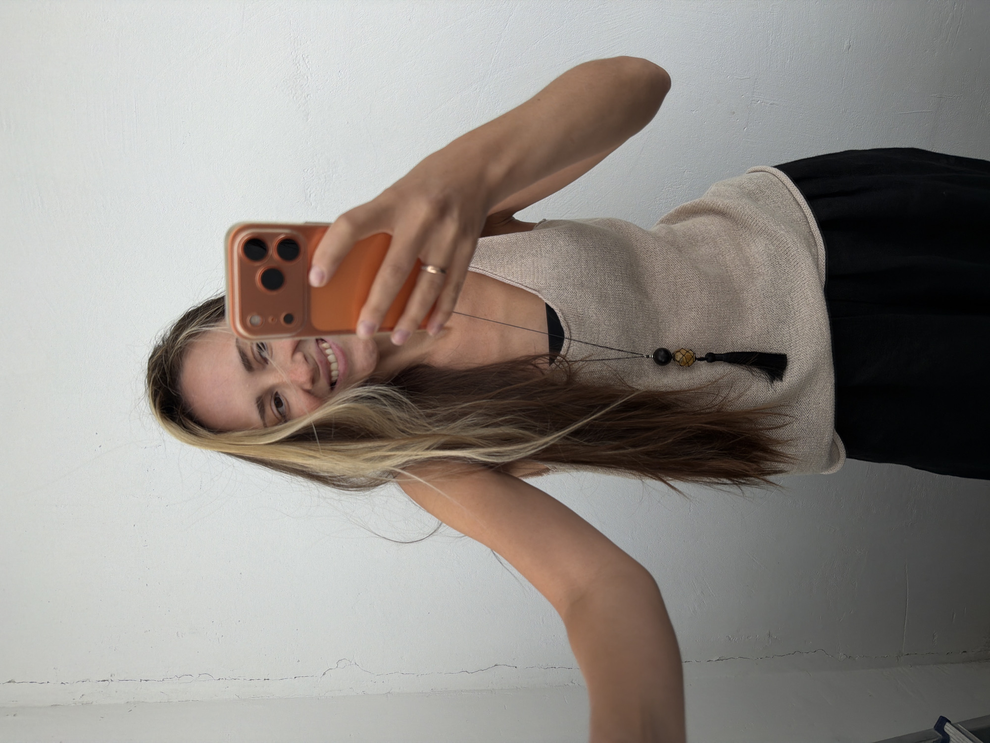 A woman with long, blonde-streaked brown hair is taking a selfie against a white, textured wall. She is wearing a light beige, sleeveless knit top over a black tank top and black wide-leg pants. A long necklace with wooden beads and a black tassel hangs from her neck. She is smiling and holding an orange smartphone in her right hand. Her left arm is extended outwards, partially out of frame. The wall has a visible crack running vertically on the left side. The lighting suggests an indoor setting. The scene appears to be in an apartment in Khimki, Russia.