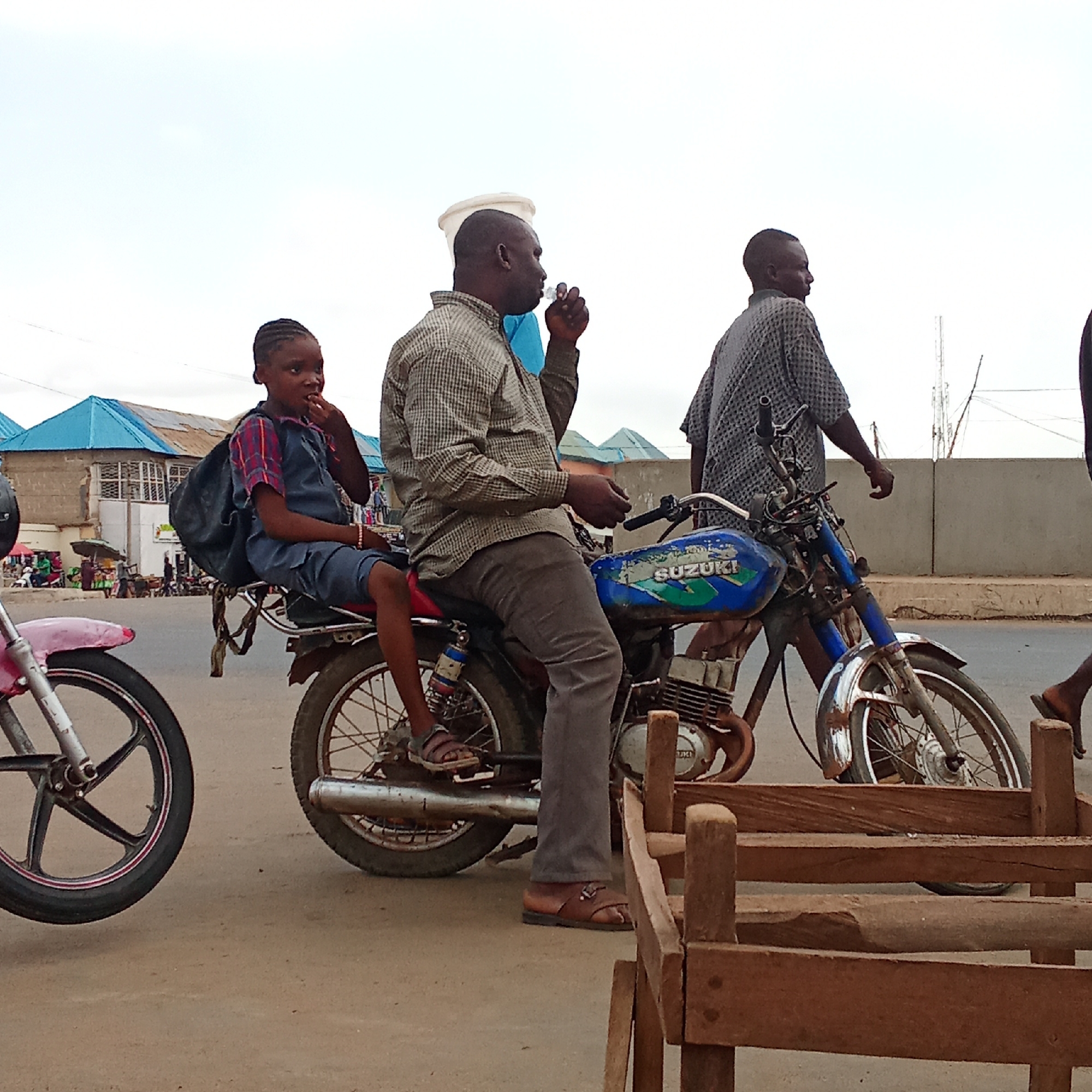 In Akwanga, Nigeria, a medium shot captures a scene on a city street. In the foreground, a pink motorcycle is partially visible on the left, and wooden planks are present on the right. A blue Suzuki motorcycle is centrally positioned, with an adult male sitting on it, holding something to his mouth. Behind him, a girl with a backpack sits on the back of the motorcycle. A man walks behind the Suzuki motorcycle, away from the viewer. A building with a blue roof is in the background, along with other people and structures.